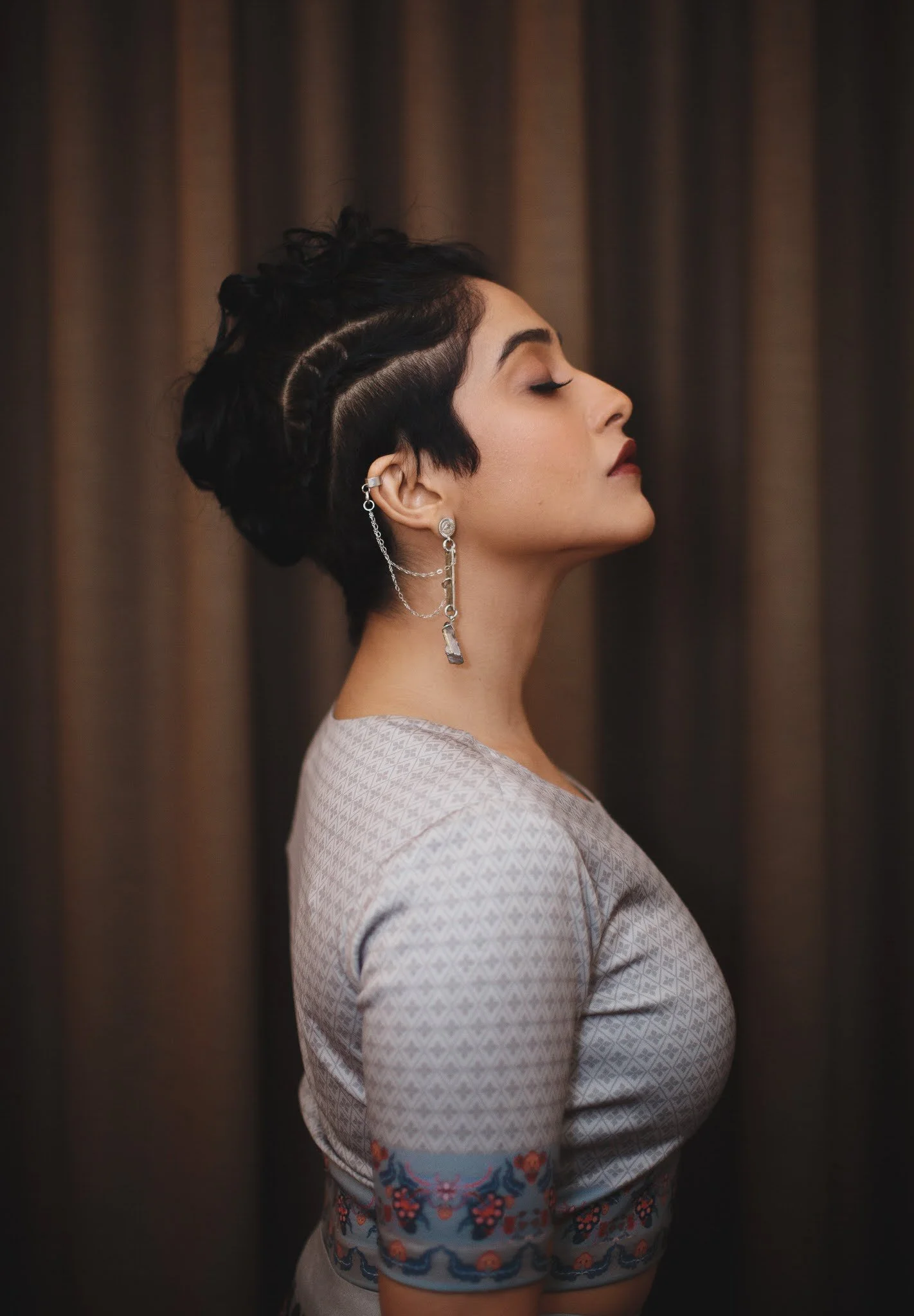 A woman with short dark hair and earrings, standing in profile with eyes closed against a dark curtain background.