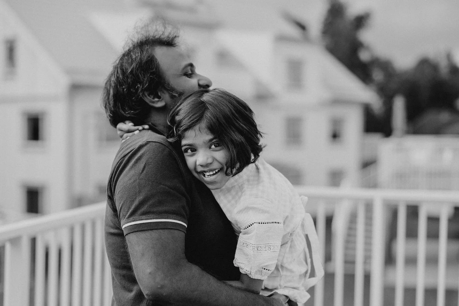 A person holding a young girl, both smiling and hugging each other on a balcony with houses and trees in the background.