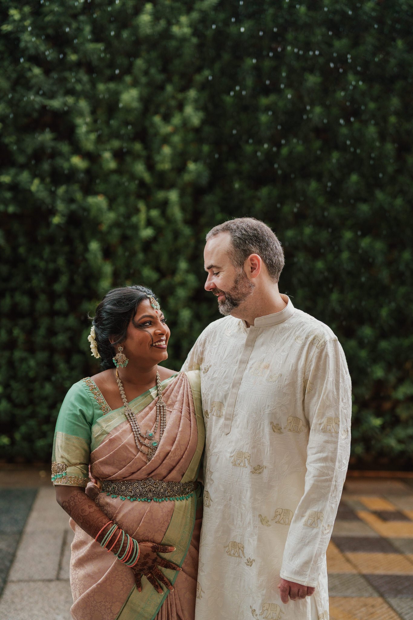 A couple dressed in traditional Indian attire is smiling and looking into each other's eyes outdoors against a green leafy background.