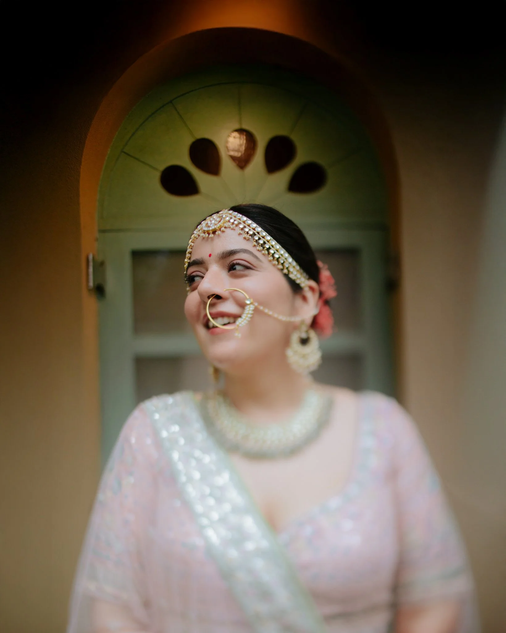 A joyful bride in traditional Indian attire, adorned with jewelry, smiling in front of an ornate arched window.
