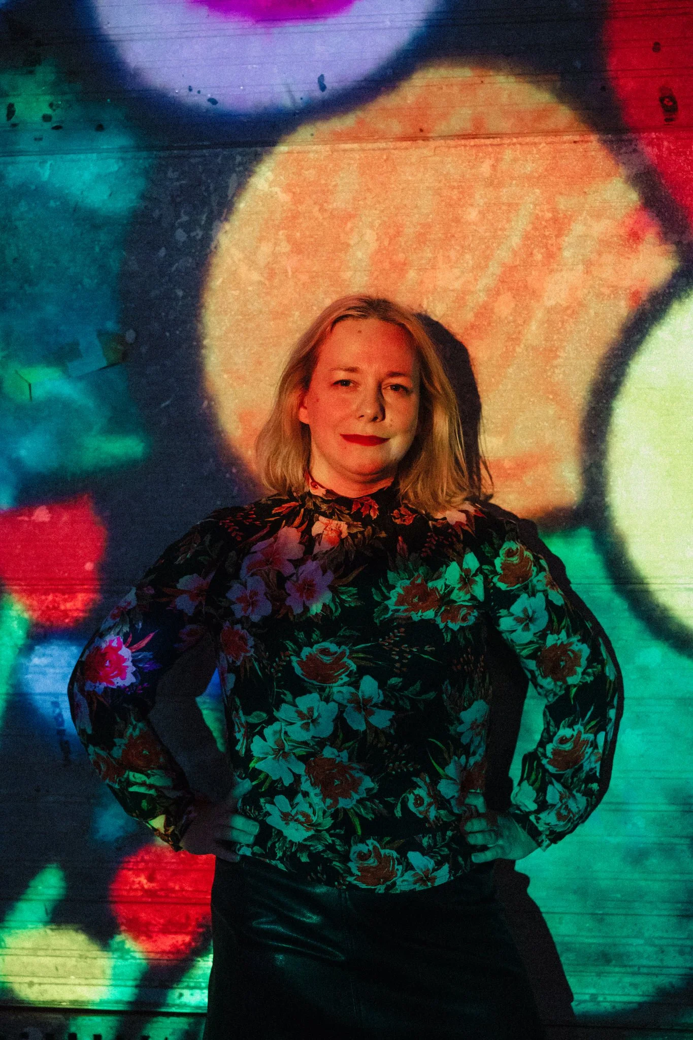 A woman with blonde hair wearing a floral long-sleeve top, standing with hands on hips in front of a colorful, abstract, illuminated background.