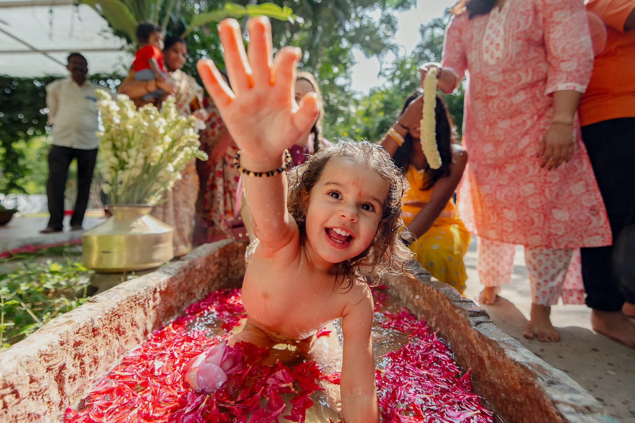 A young girl with curly hair, smiling and waving, sitting in a rectangular water trough filled with flower petals. She is showered with water and surrounded by people, some dressed in traditional Indian clothing, during a festive celebration outdoors