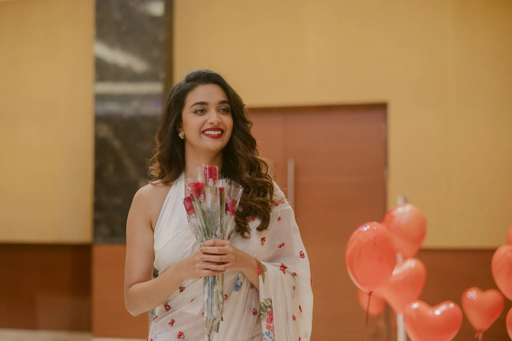 A woman with long curly hair, wearing a white saree with red floral patterns, holding a bouquet of red roses, smiling at the camera, with pink heart-shaped balloons in the foreground.