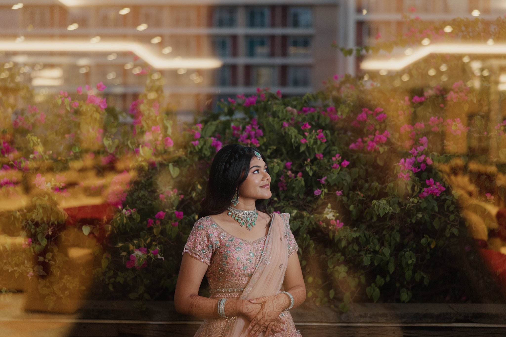 Young woman dressed in traditional Indian attire, wearing jewelry, standing by a window with pink flowers in the background, looking outside.