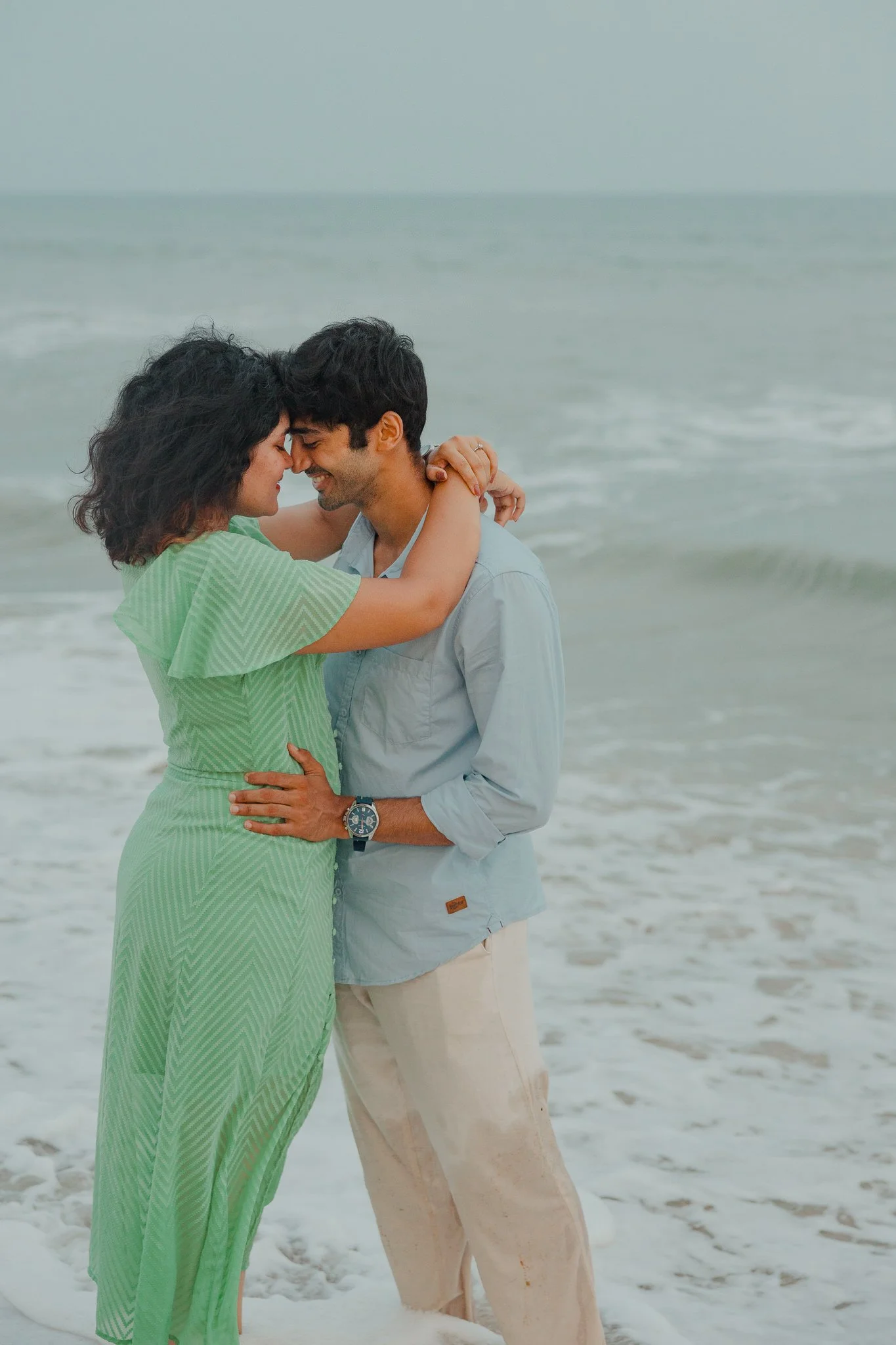 A couple embracing on the beach, with the waves in the background, smiling and touching foreheads.