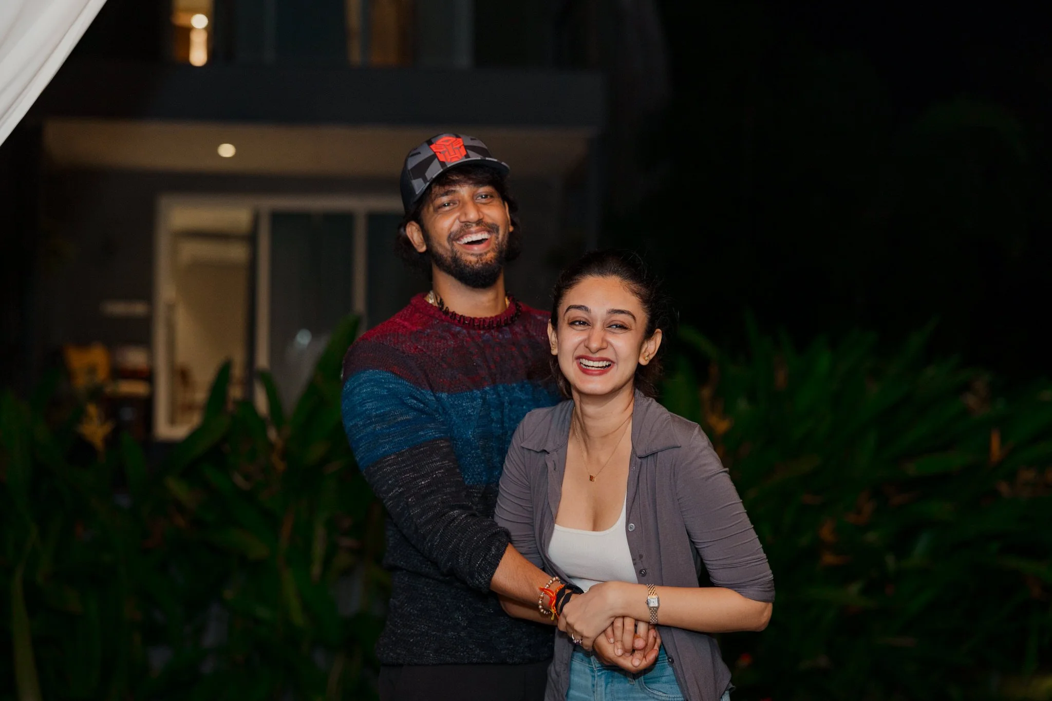 A happy young couple with dark hair and medium skin tones smiling and holding hands outdoors at night, with greenery and a modern building in the background.