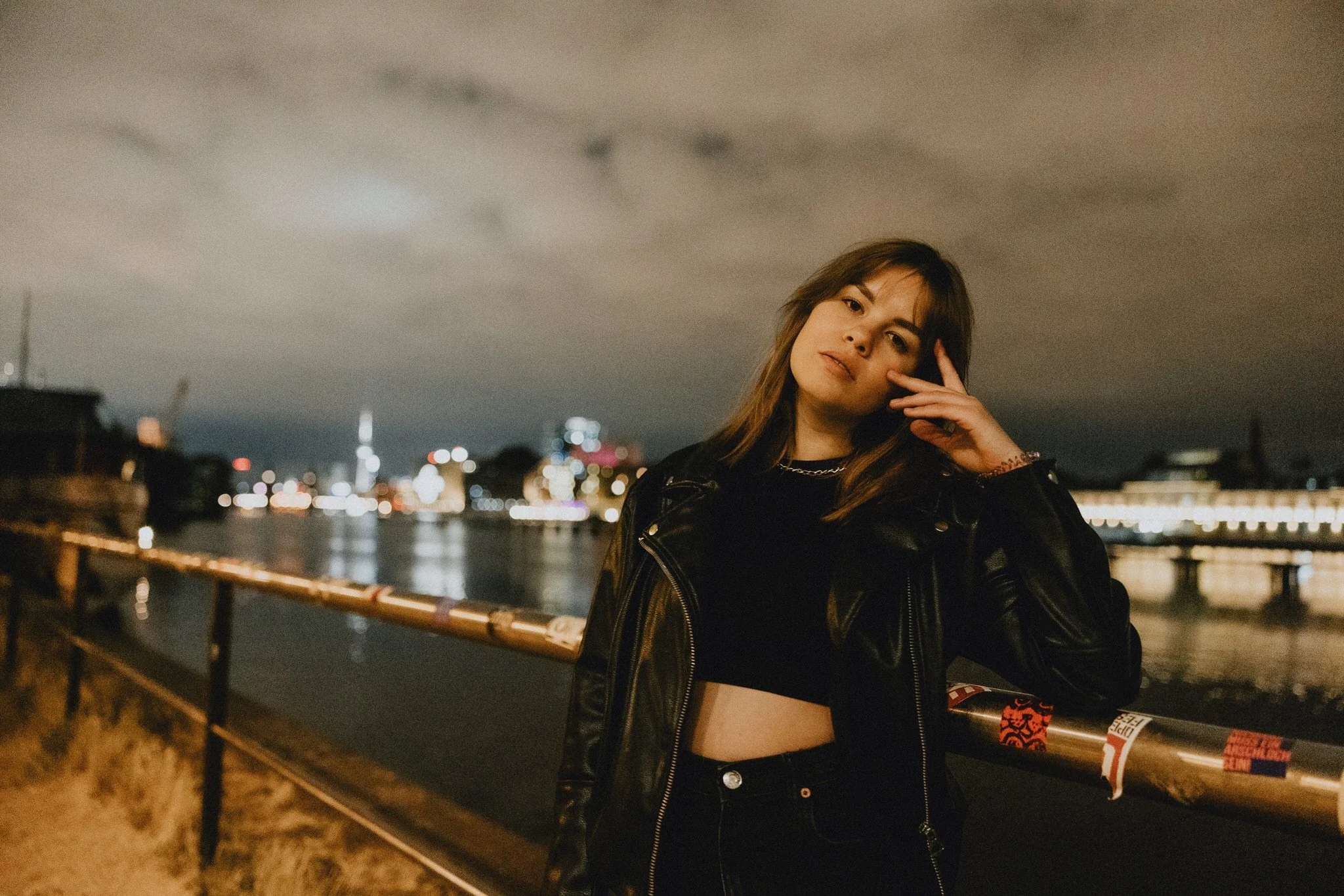 Young woman with brown hair, wearing a black leather jacket and crop top, standing near a river at night with city lights in the background.