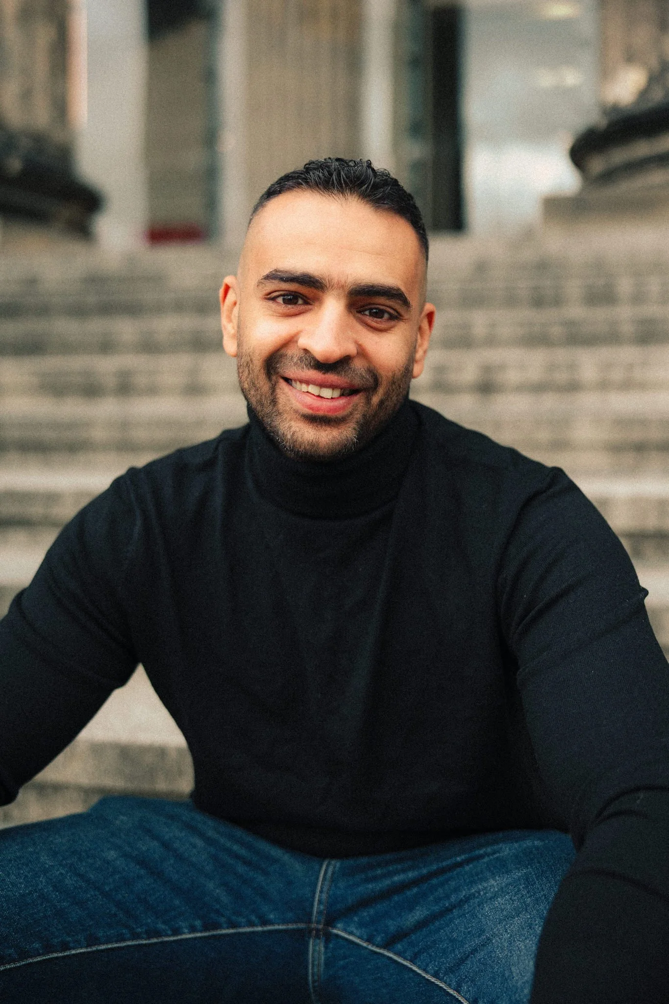 A smiling man sitting on outdoor stairs wearing a black turtleneck and jeans.