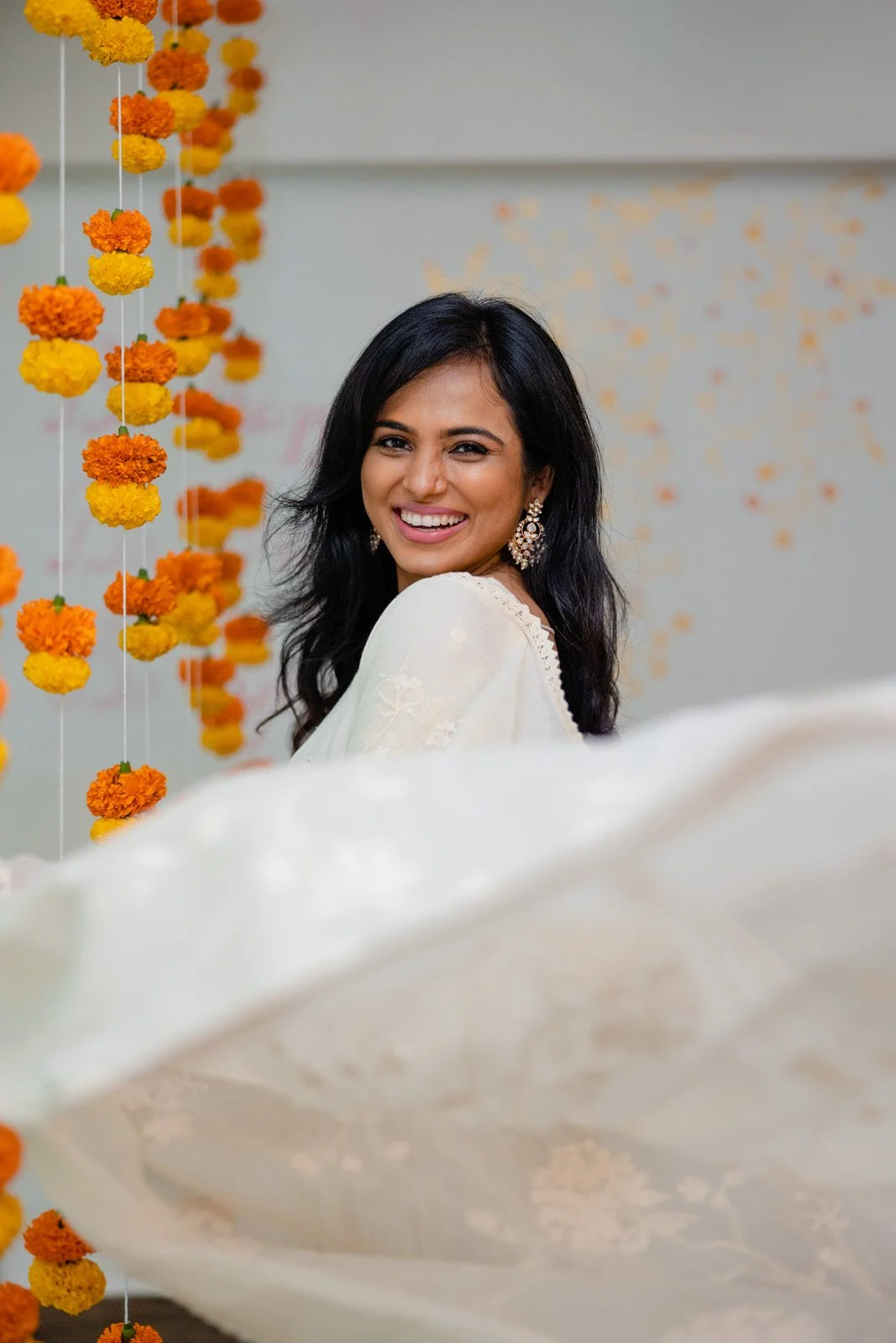 Woman smiling with dark hair and earrings, celebrating with orange and yellow marigold flower garlands, at a festive event.
