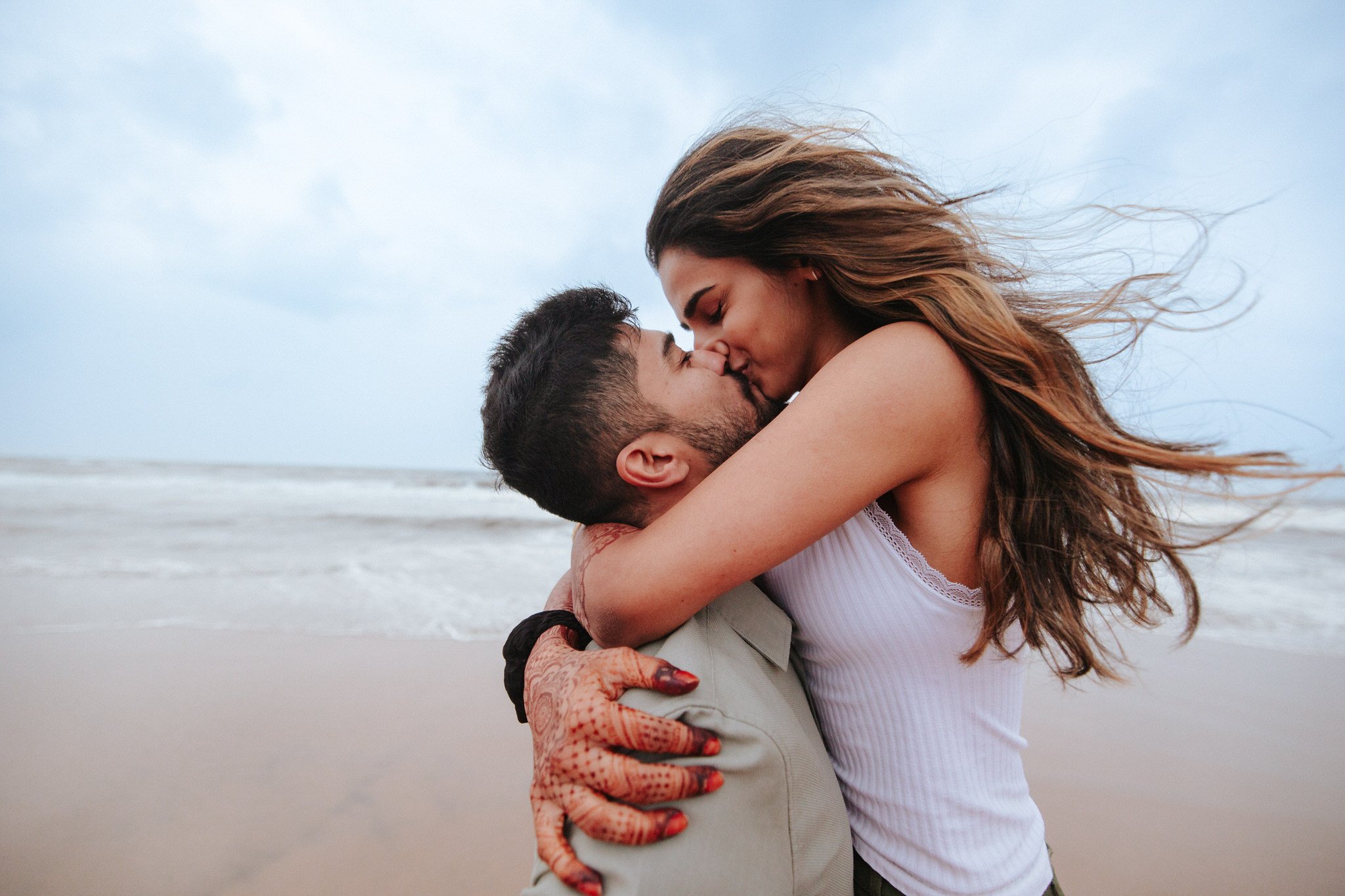 A couple kissing passionately on the beach, with wind blowing their hair over a cloudy sky.