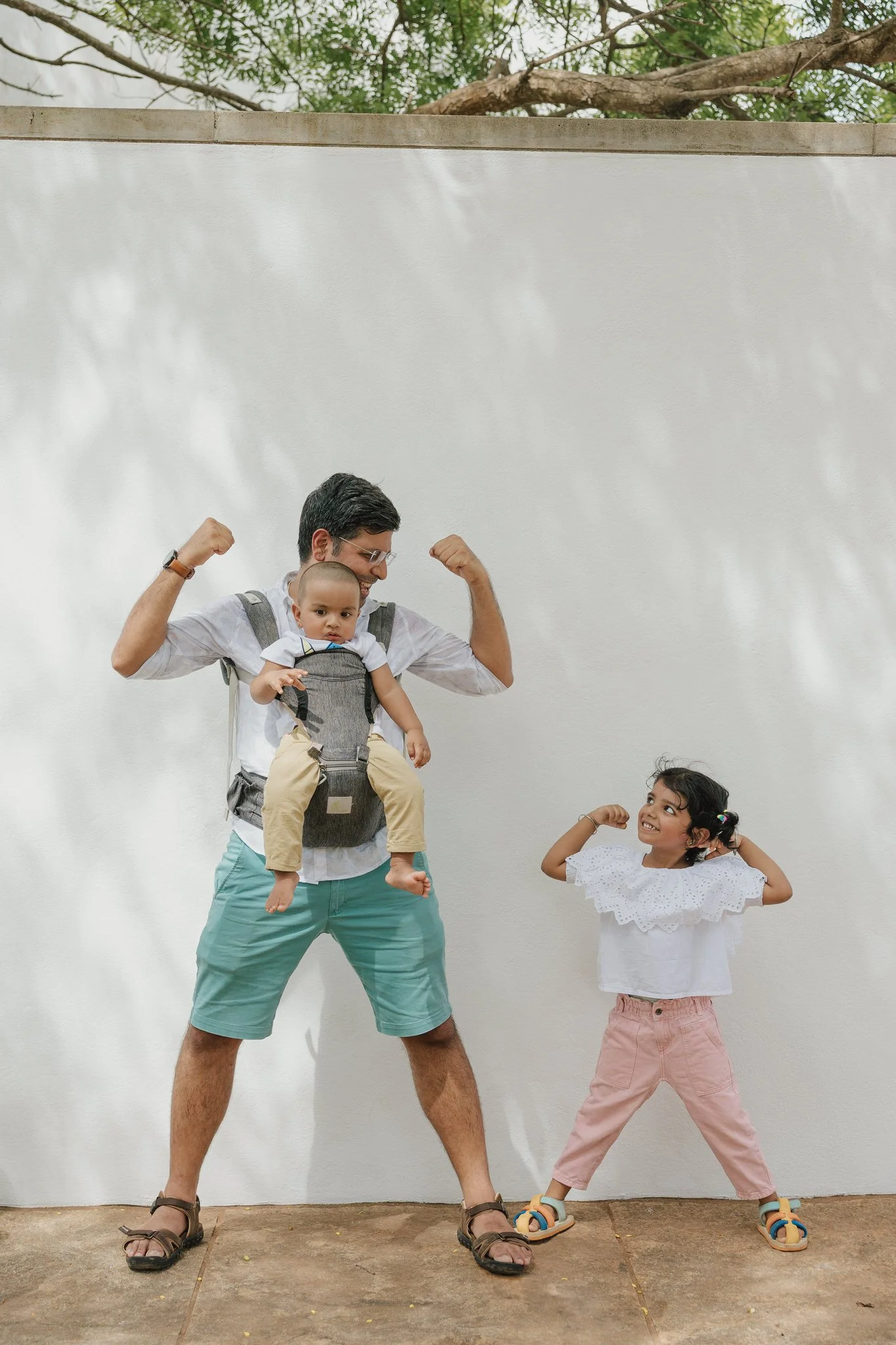 A man with glasses and a beard, wearing shorts and sandals, is standing against a white wall, flexing his arms. He has a toddler in a baby carrier on his chest, who is looking forward. A young girl with pigtails, wearing a white blouse with ruffled c