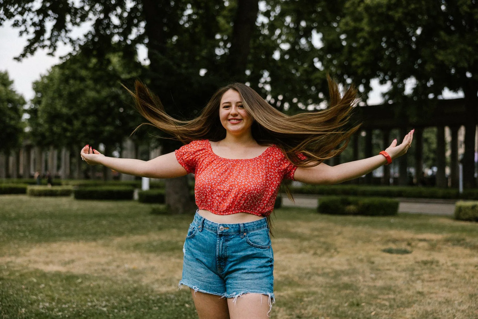 Young woman with long hair wearing a red floral crop top and blue denim shorts, standing outdoors with arms outstretched and smiling