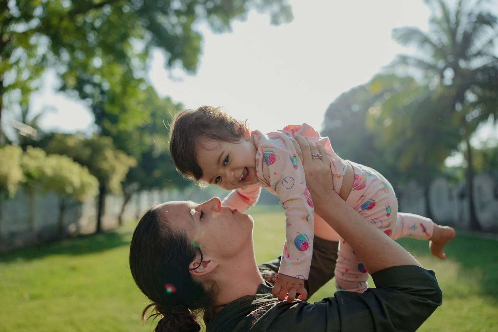 A woman with dark hair in a ponytail lifts a smiling young girl with dark hair and pajamas into the air outside in a park on a sunny day.