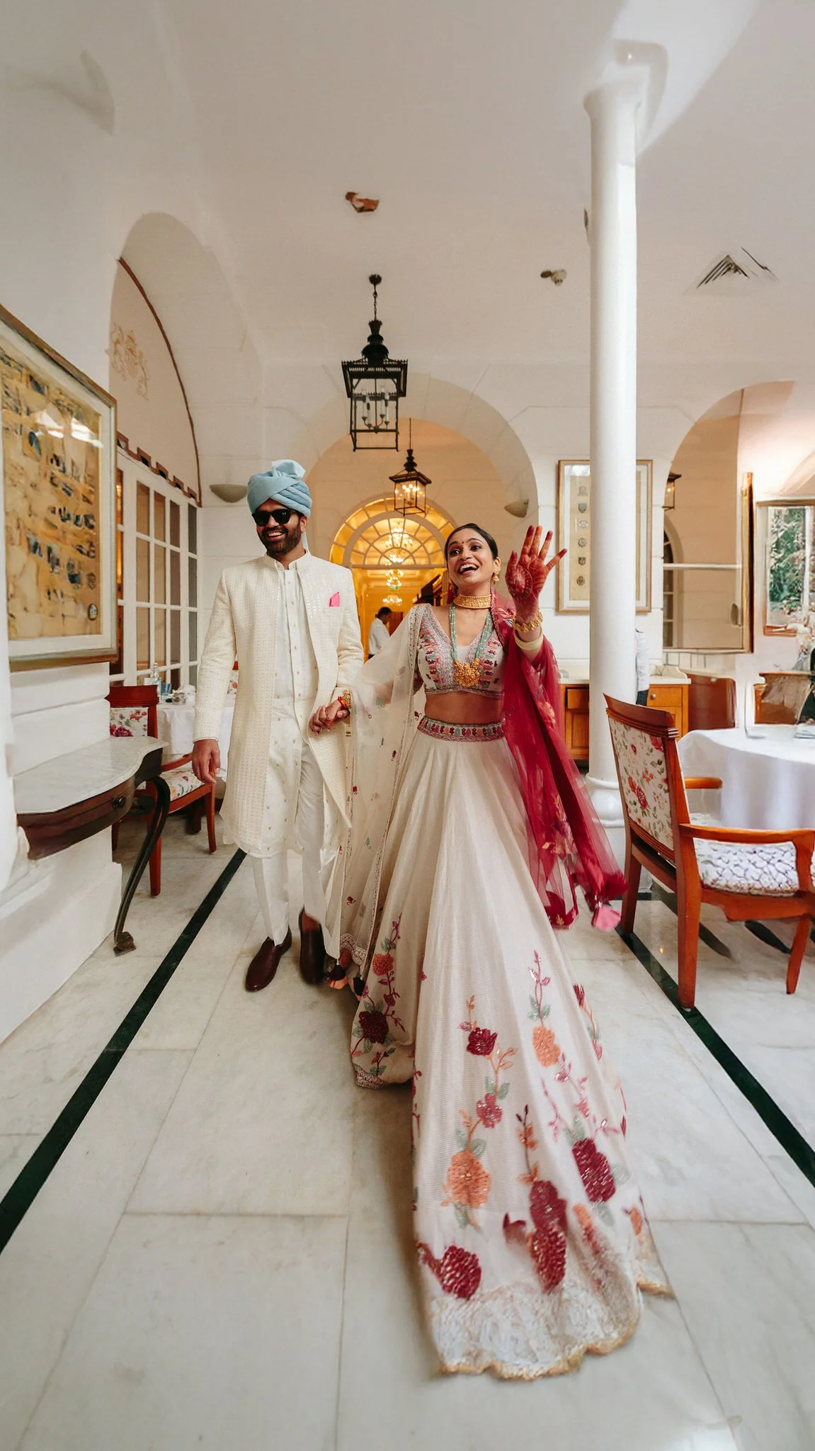 A bride and groom walking inside a decorated venue, with the bride waving and smiling. The bride wears a white embroidered lehenga and jewelry; the groom wears traditional Indian attire with a turban and sunglasses.