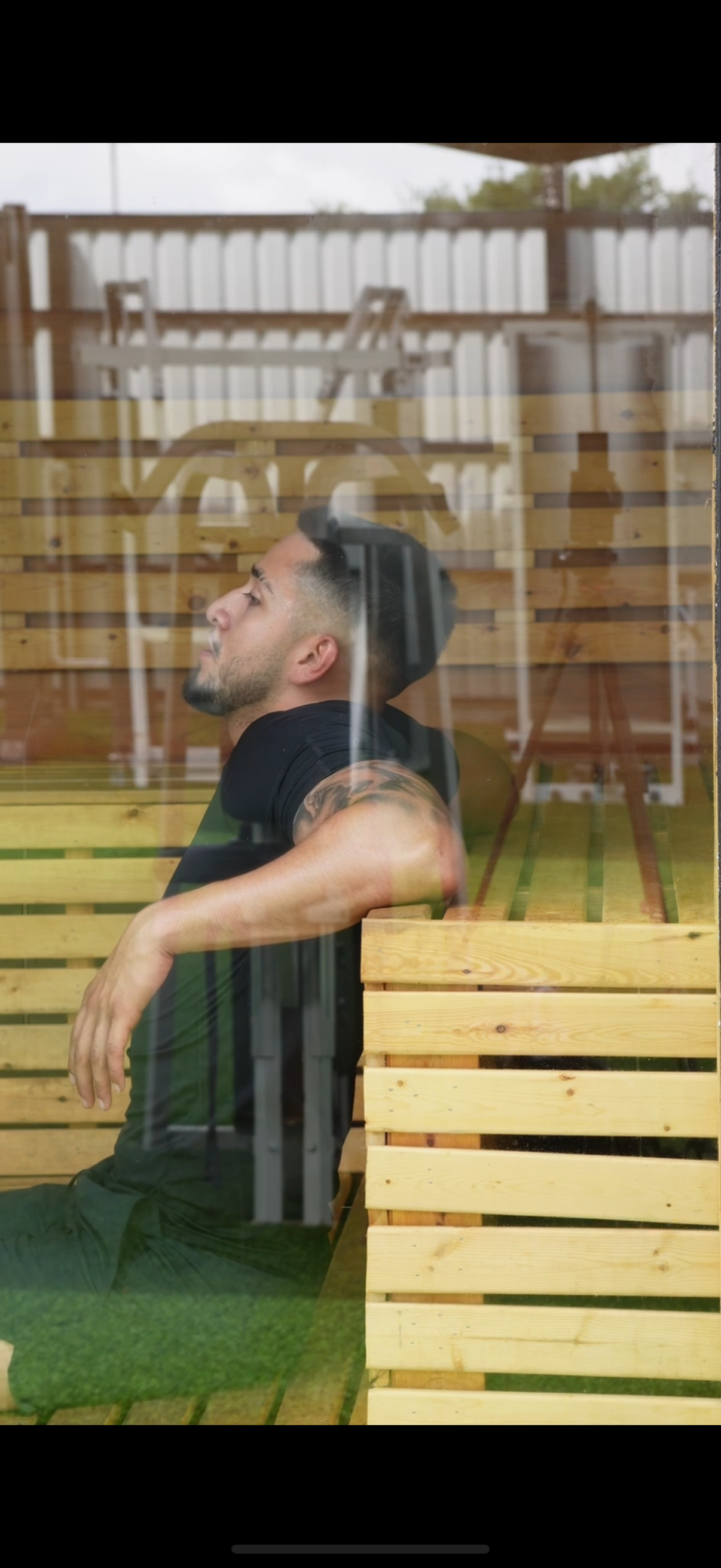 A man with a tattoo on his right arm, short dark hair, and a beard, sitting and leaning back with his arms resting on a wooden bench inside a sauna or wooden structure, seen through a glass window with reflections.