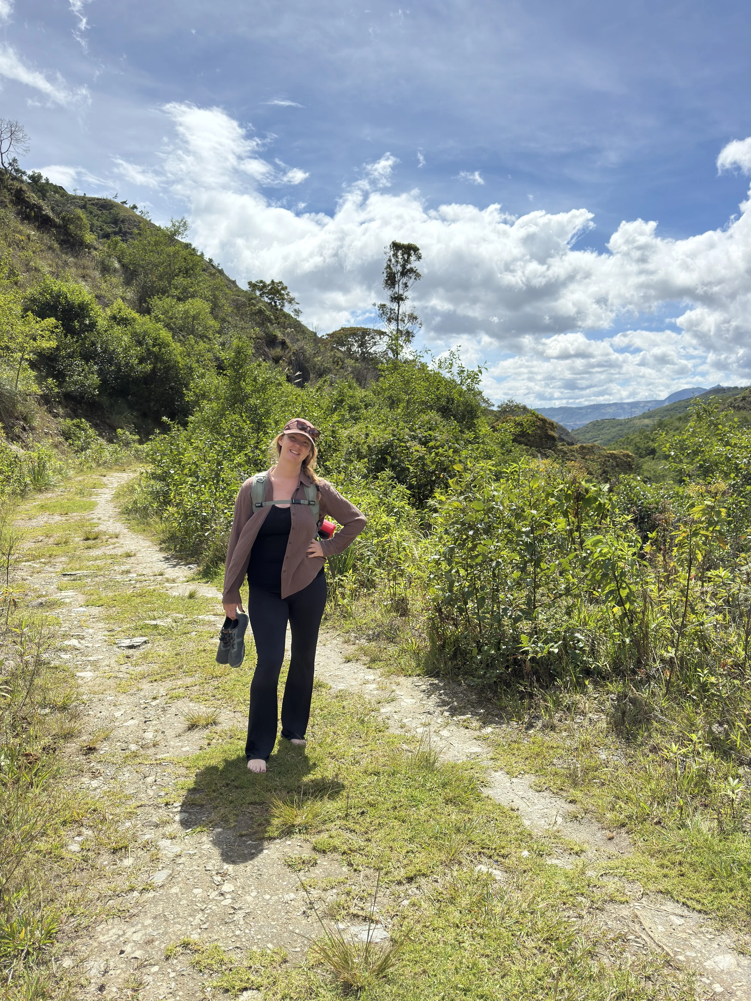 A woman hiking on a dirt trail in a lush green canyon, with mountains and a partly cloudy sky in the background.