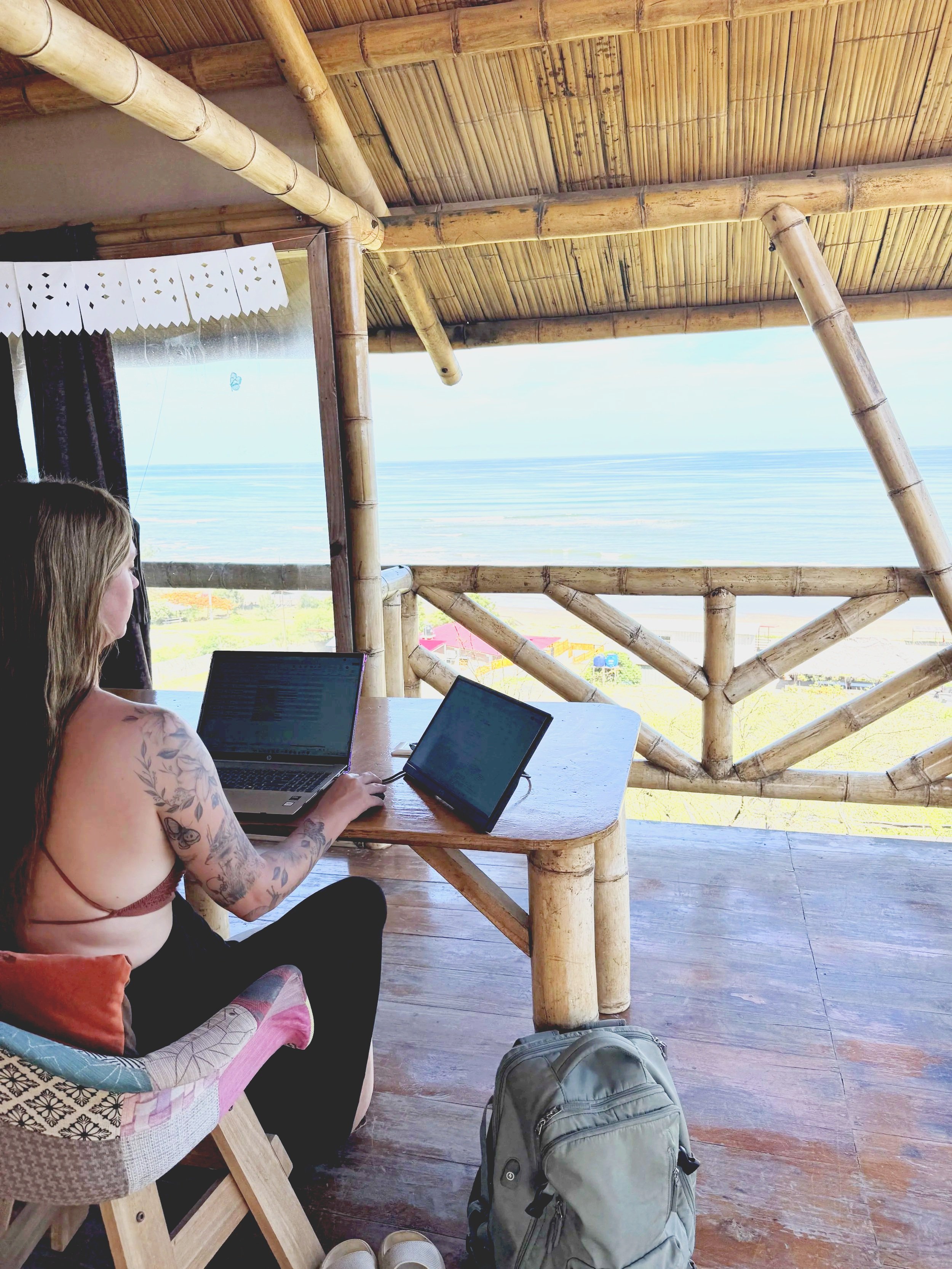 Woman with tattoos sitting at a wooden table on a bamboo balcony, working on a laptop and tablet, overlooking a beach and ocean.
