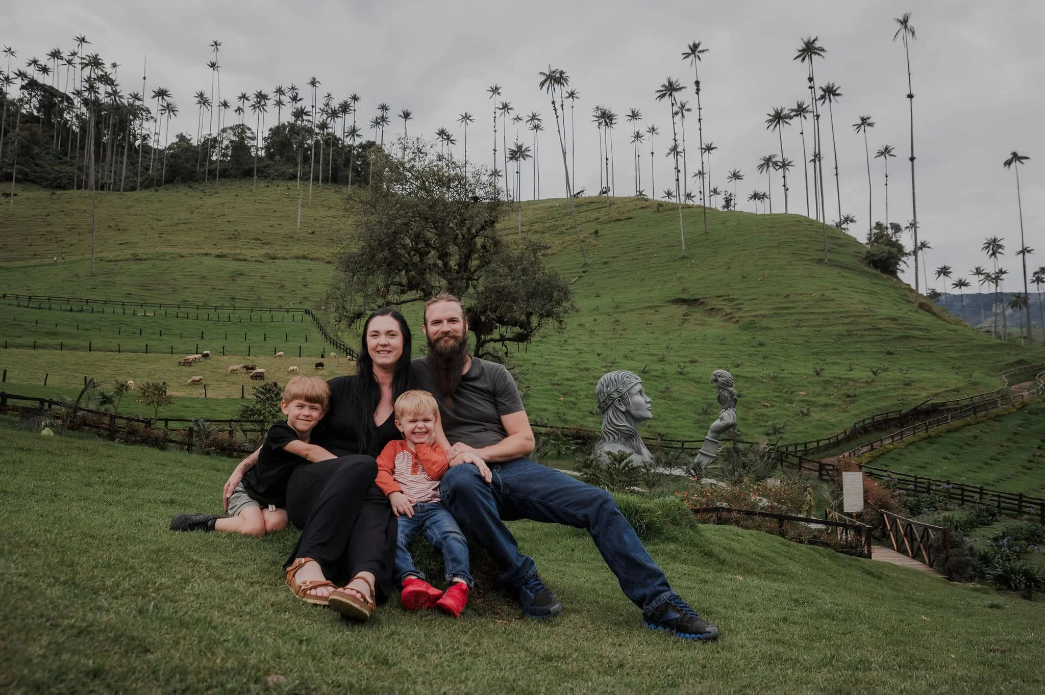 A family of four smiling and sitting on the grass in front of a lush, green hillside with tall palm trees and sculptures, under a cloudy sky.