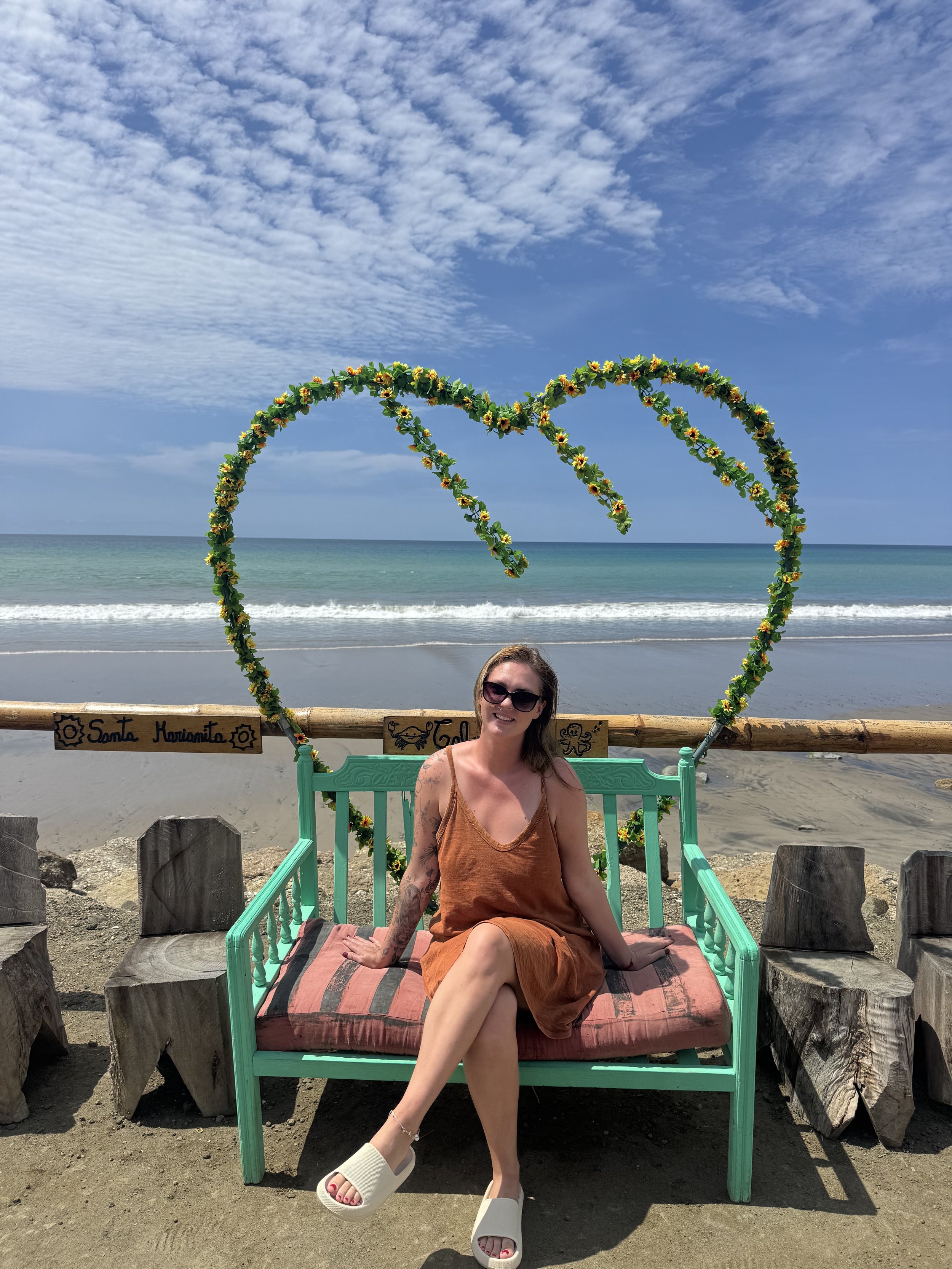A woman in sunglasses and a brown dress sitting on a green bench on a beach, with a heart-shaped floral decoration behind her and the ocean in the background.