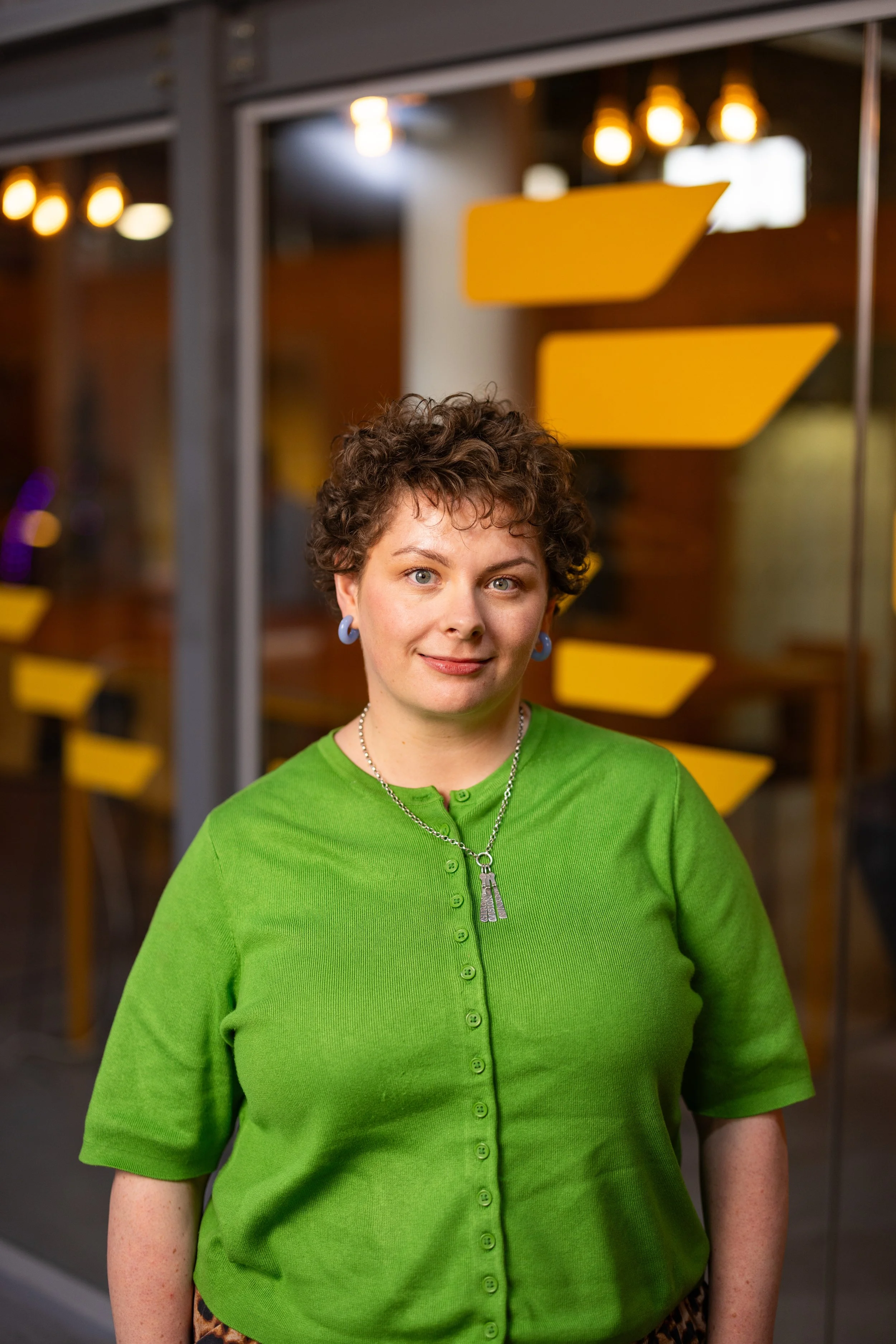 A woman with short, curly brown hair wearing a bright green cardigan with buttons, a necklace with a key pendant, and earrings, standing indoors with blurred yellow lights and signs in the background.