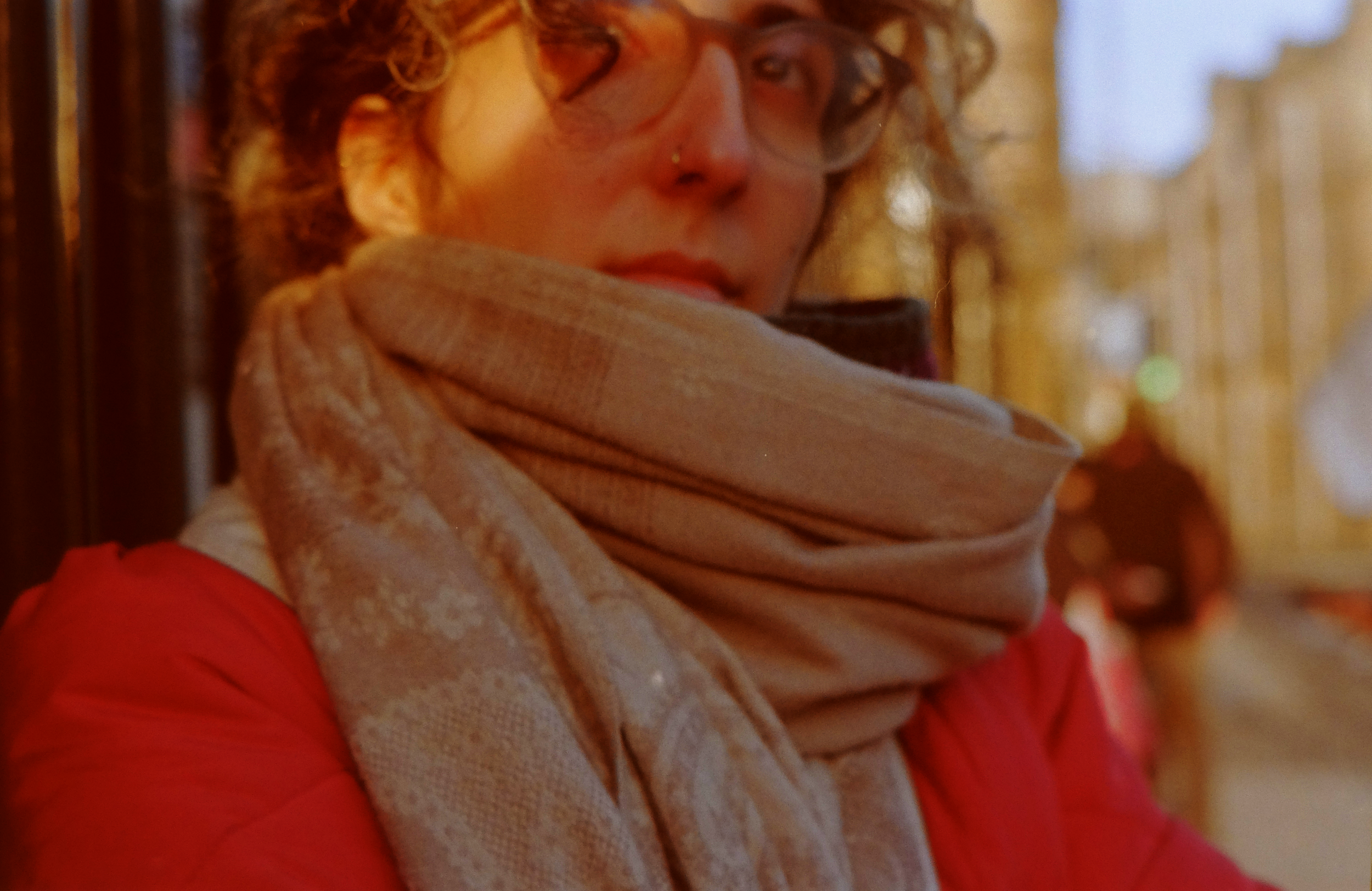 A woman with curly hair, glasses, and a nose piercing wearing a beige scarf and red jacket, standing outdoors with blurred background.