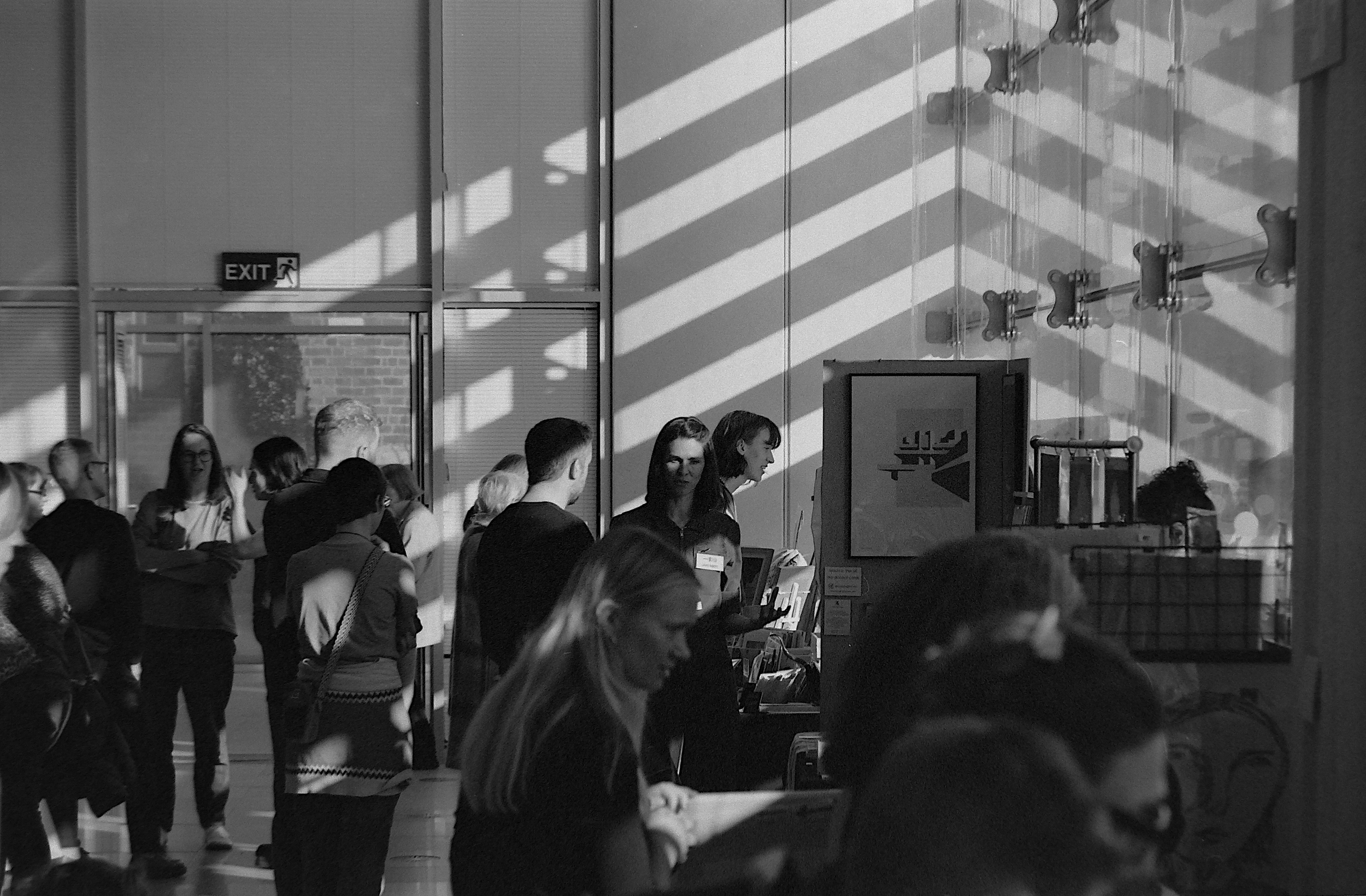 People waiting in line inside a modern building with glass walls, sunlight casting striped shadows, an exit sign, and a kiosk or information booth.