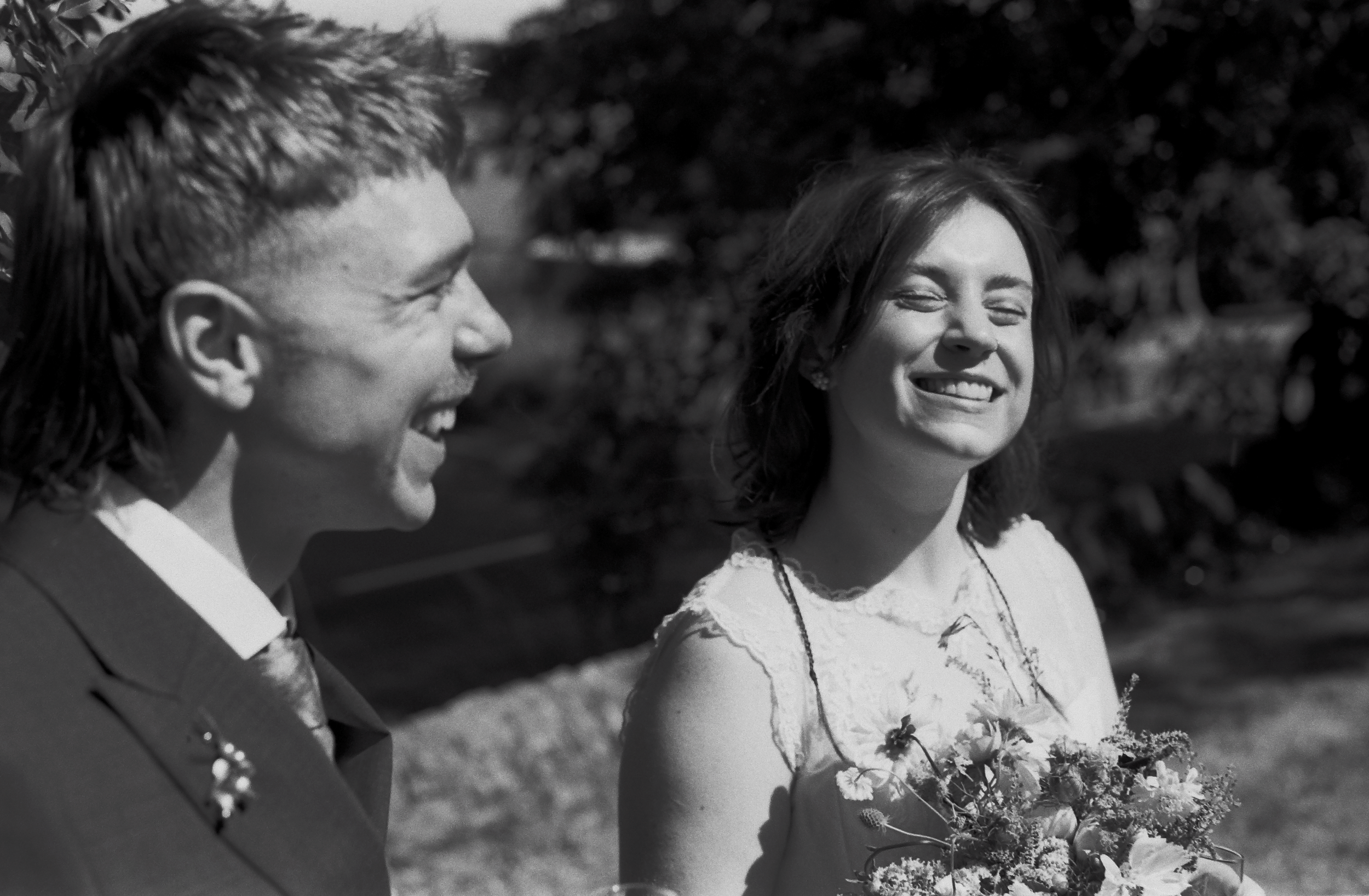 A man and a woman are outdoors, smiling and looking at each other. The woman is holding a bouquet of flowers. The man is wearing a suit, and the woman is dressed in a floral dress. They appear happy and content.