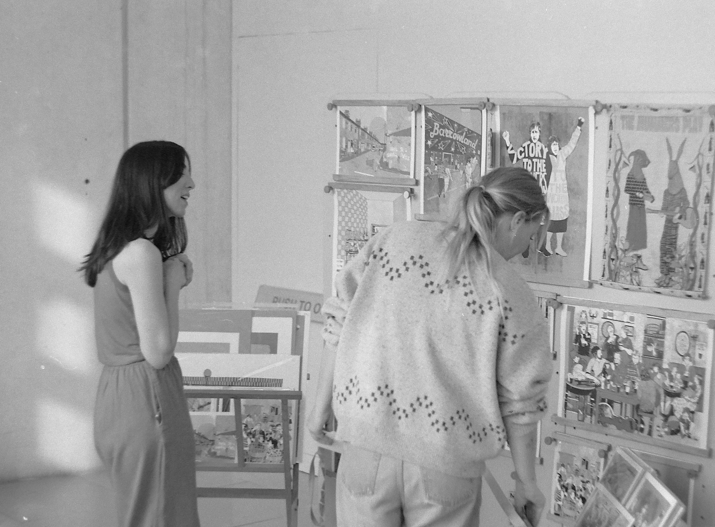 Two women viewing art on display in an art gallery, one smiling and the other leaning forward to examine the artwork.