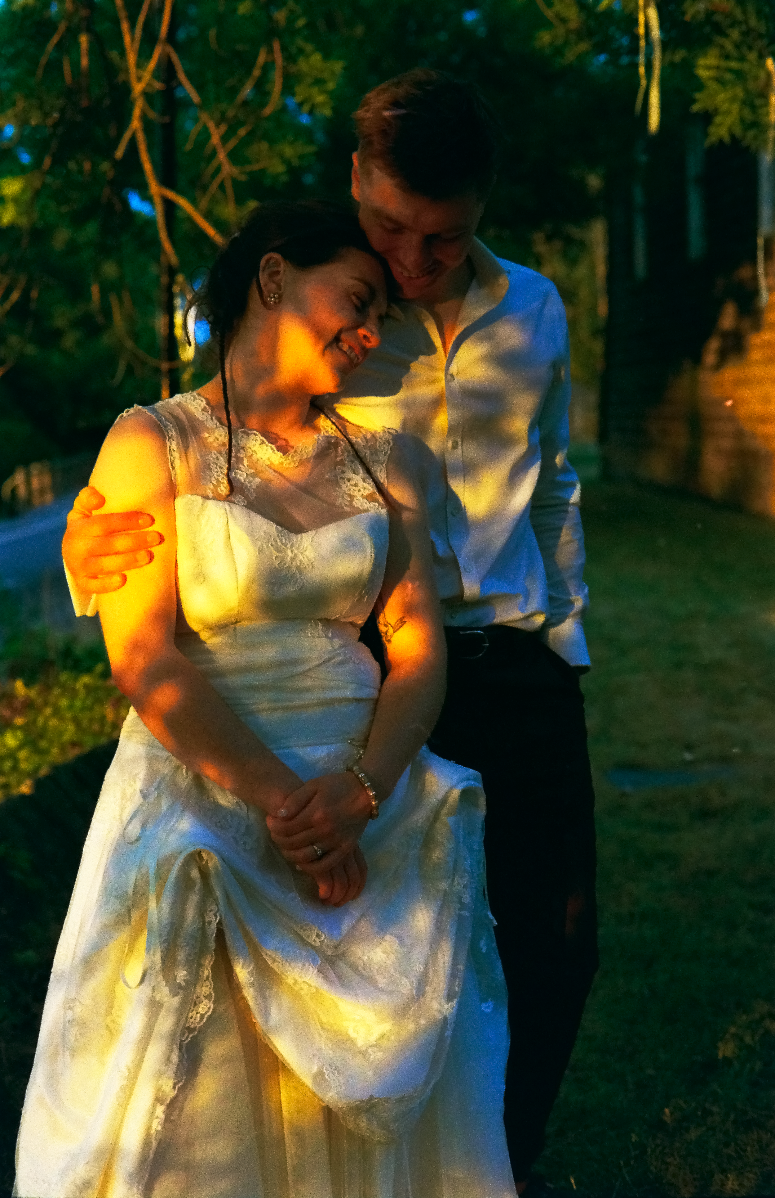 A couple dressed in wedding attire smiling and embracing outdoors at night, with trees and colorful lighting in the background.
