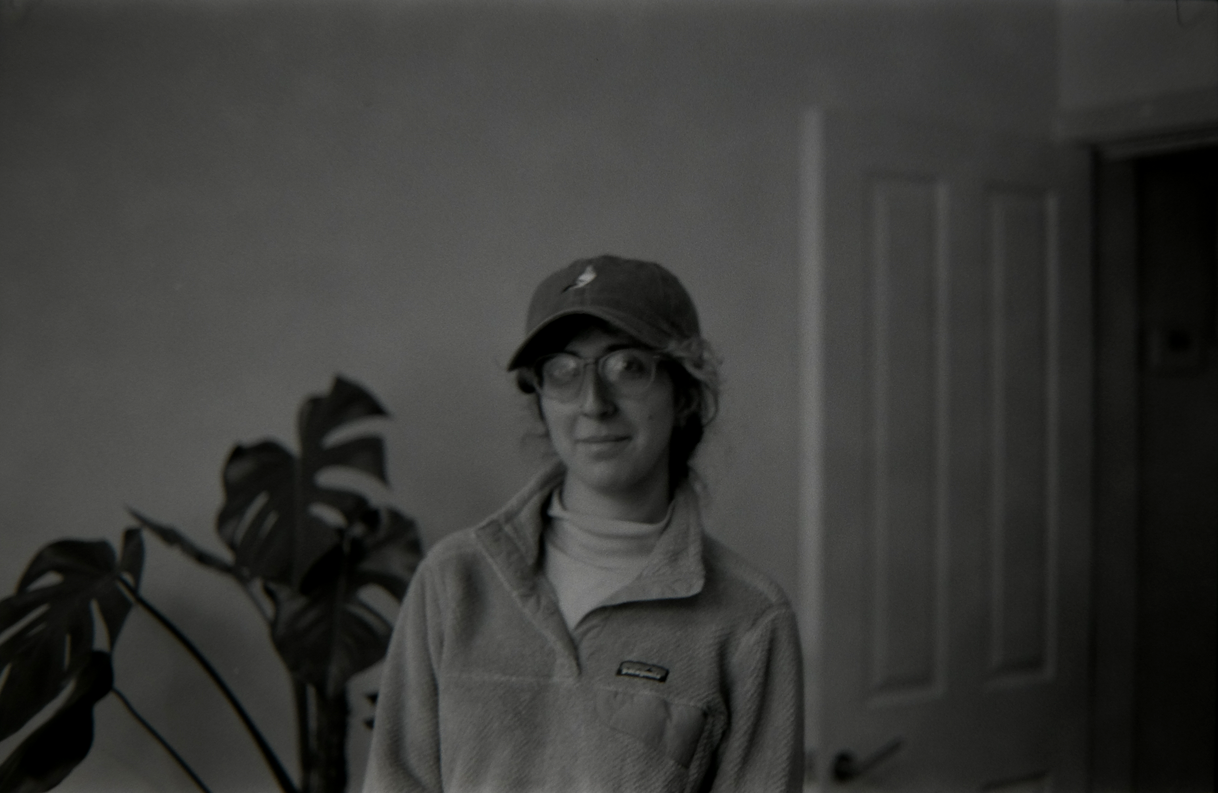 A woman wearing glasses, a baseball cap, and a fleece jacket standing indoors near a plant and a door.