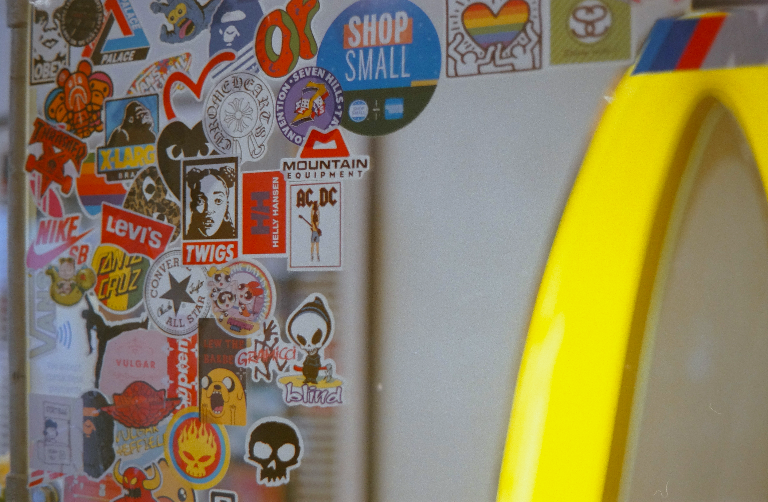 Close-up of a grey surface covered with various colorful stickers, including brands like Nike, Levi's, Converse, and Marvel characters, with part of a bright yellow object on the right.