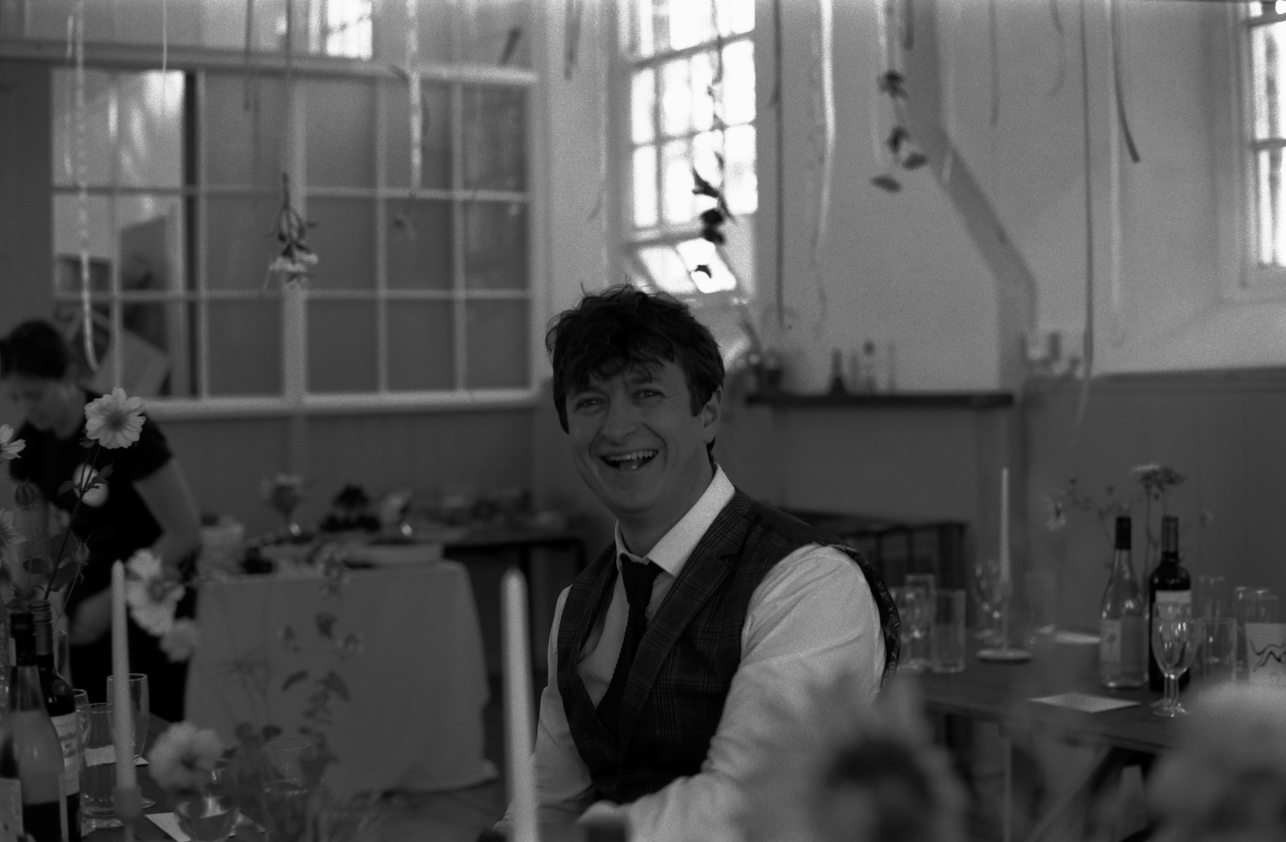 A man with dark, tousled hair and a wide smile is sitting at a table in a bright room with large windows. The room has various bottles and flowers, suggesting a celebration or gathering. In the background, there is a person with glasses and short hai