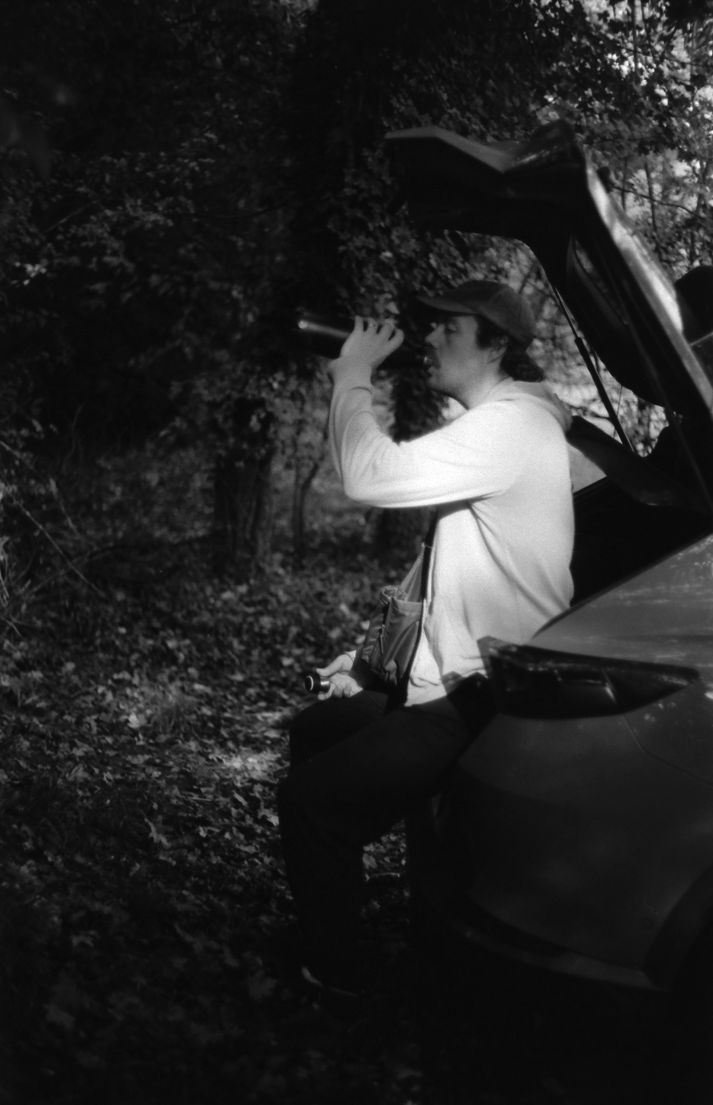 A person with long hair, wearing a hat and a light-colored jacket, sitting on the edge of a vehicle in a wooded area, drinking from a bottle.