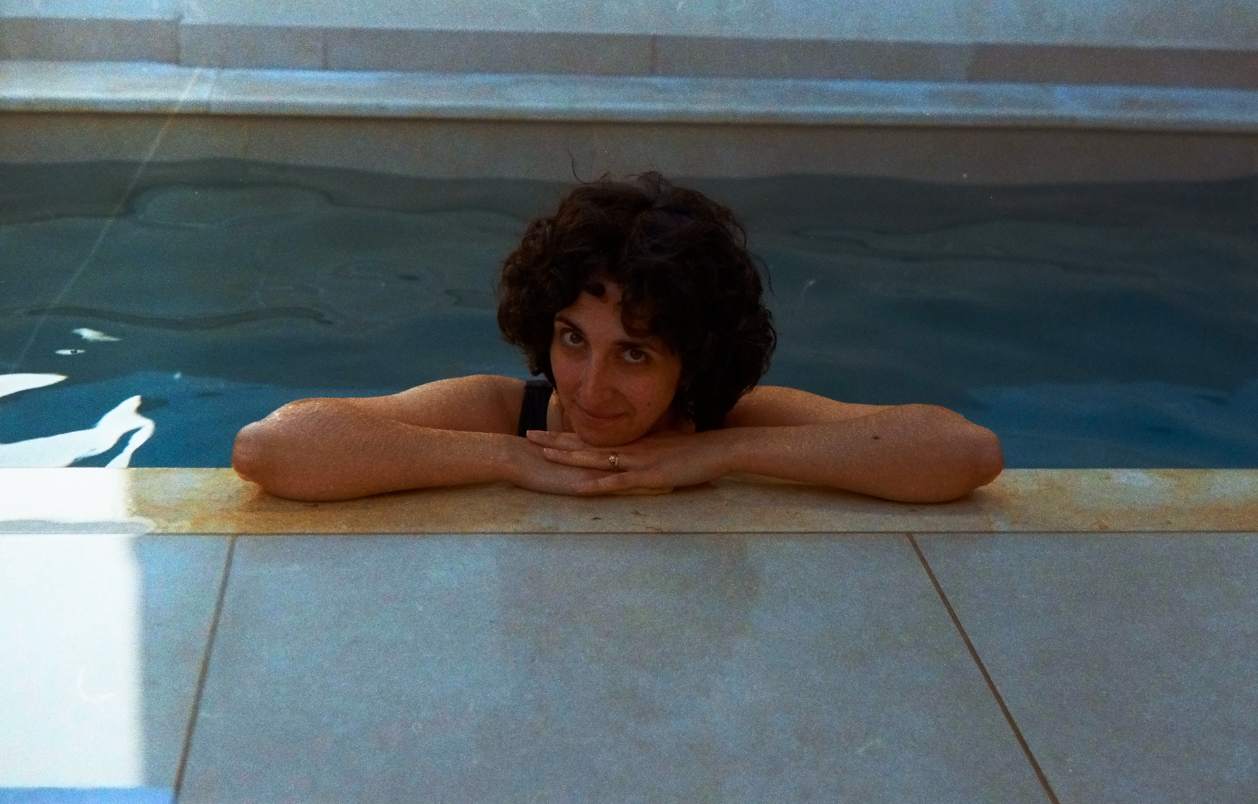 Woman with curly hair resting her chin on her crossed arms at the edge of a swimming pool.