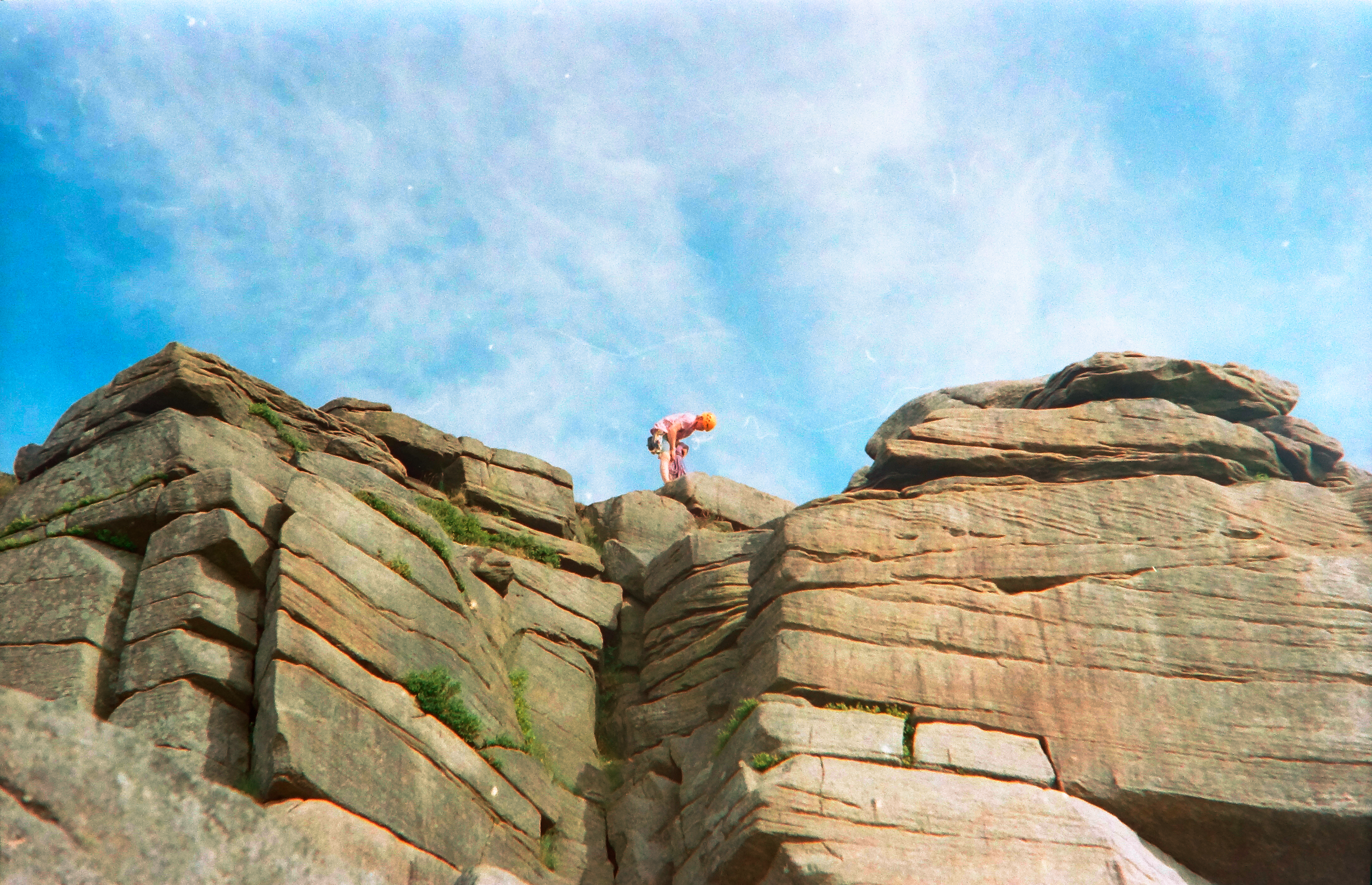 Person wearing an orange helmet and pink outfit climbing on large rock formations against a bright blue sky.