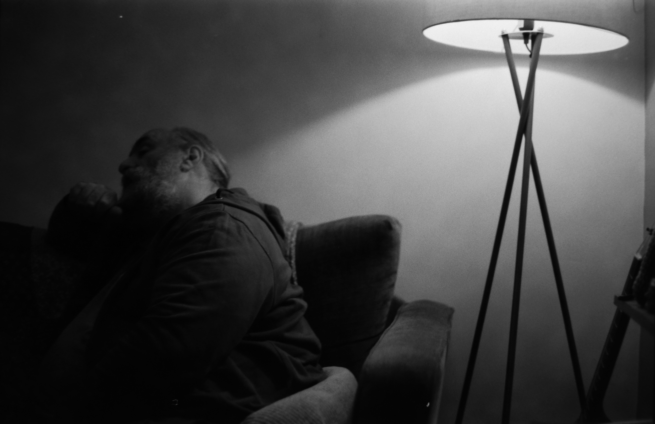A black and white photograph of an older man with a beard, sitting on a sofa with his eyes closed, resting his head on a cushion. A tall, modern floor lamp is turned on to his right, illuminating the scene.