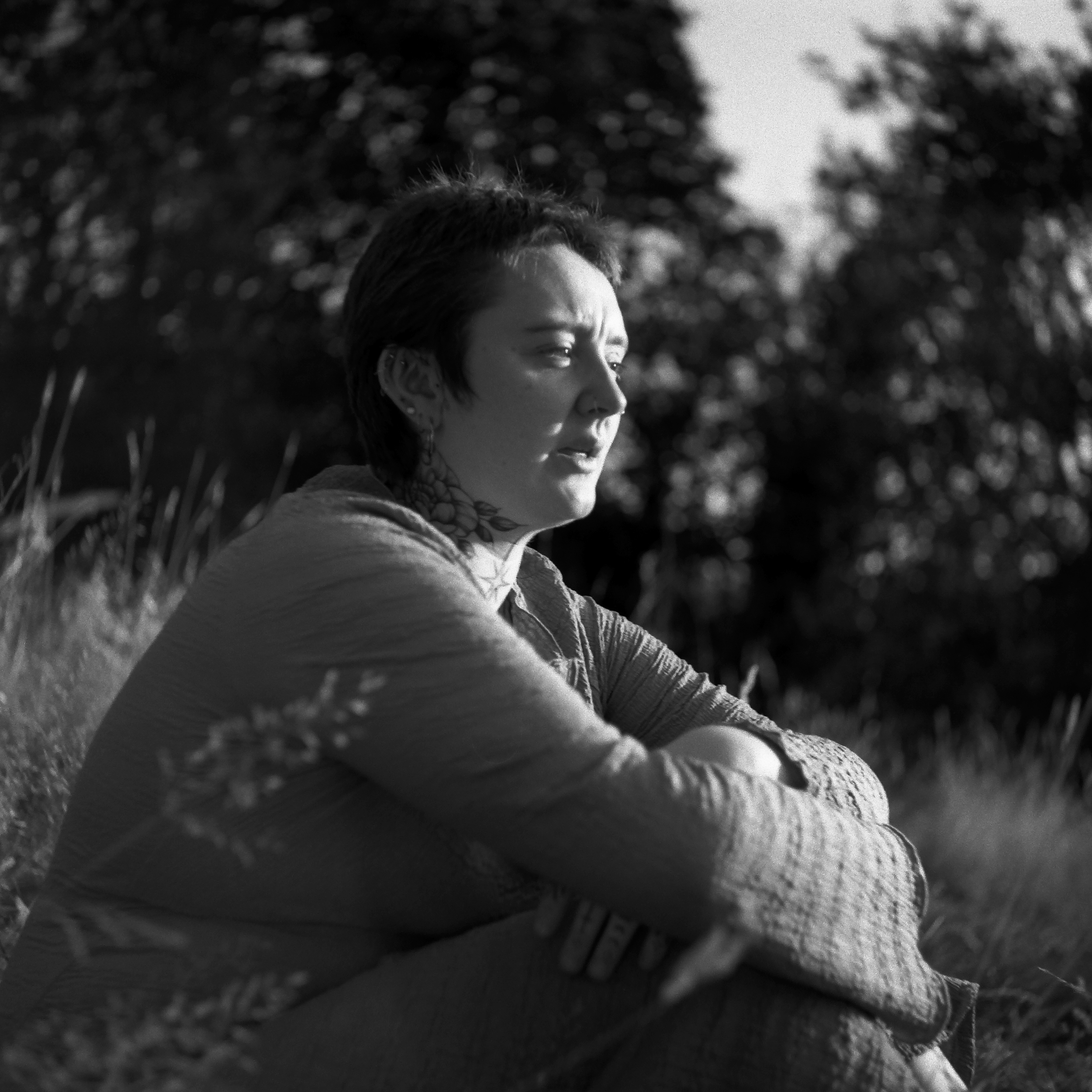 Close-up of a person's face with short hair, looking to the side, with a background of trees and bokeh effect in black and white.