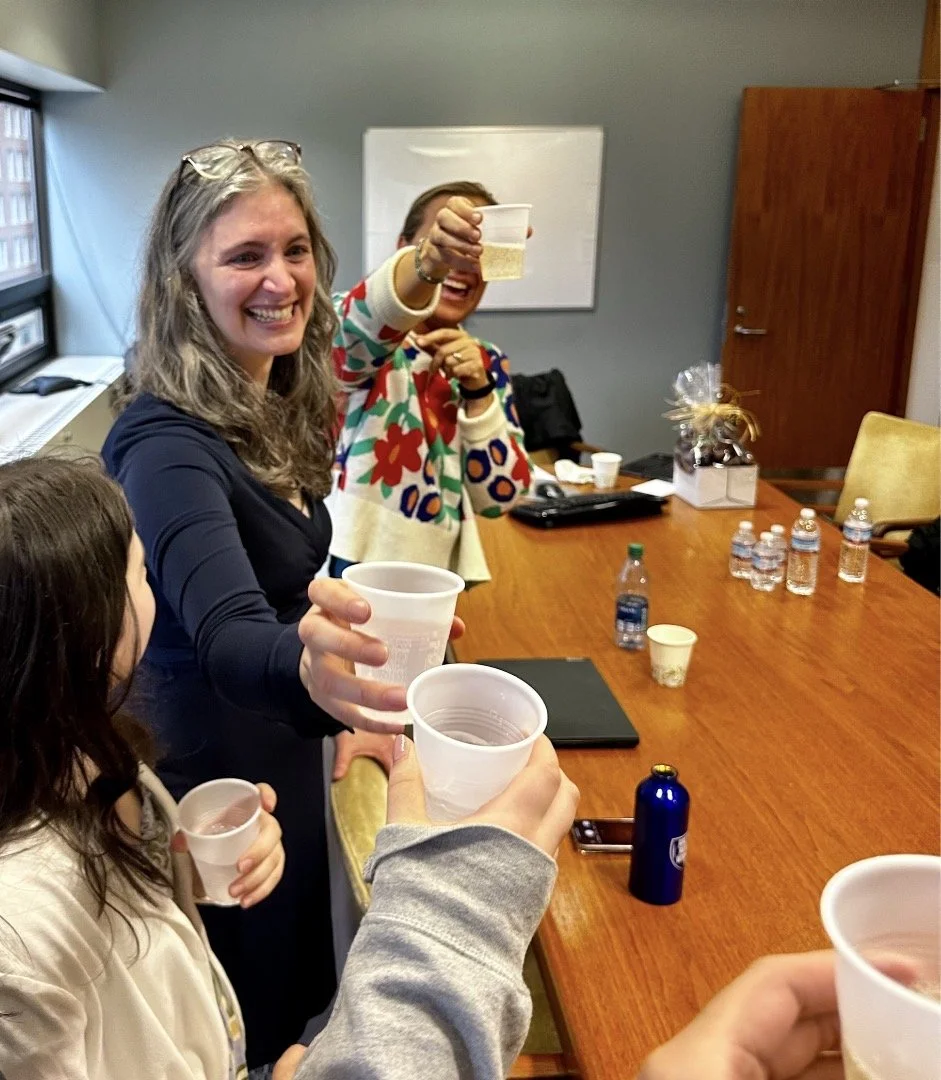 Woman smiling widely, toasting with bubbly water in plastics cups with others around a large wooden table.