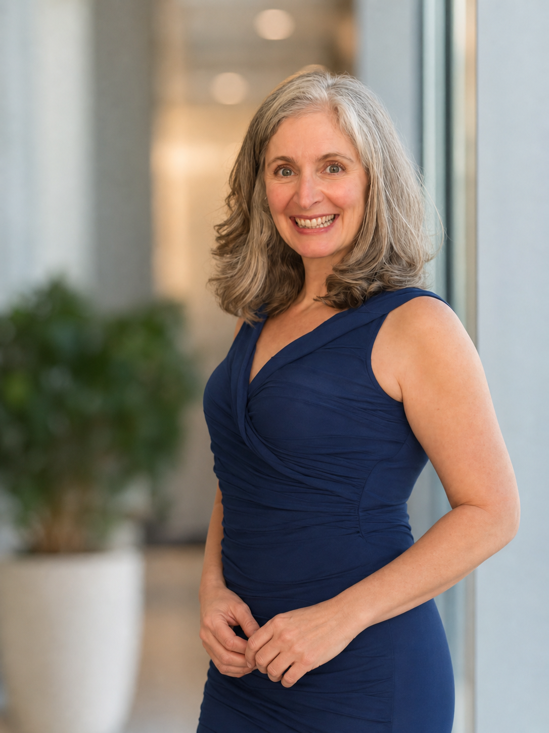 Dr. Bridget Colacchio, a smiling woman with gray hair wearing a sleeveless blue dress, standing indoors near large windows with a blurred background.