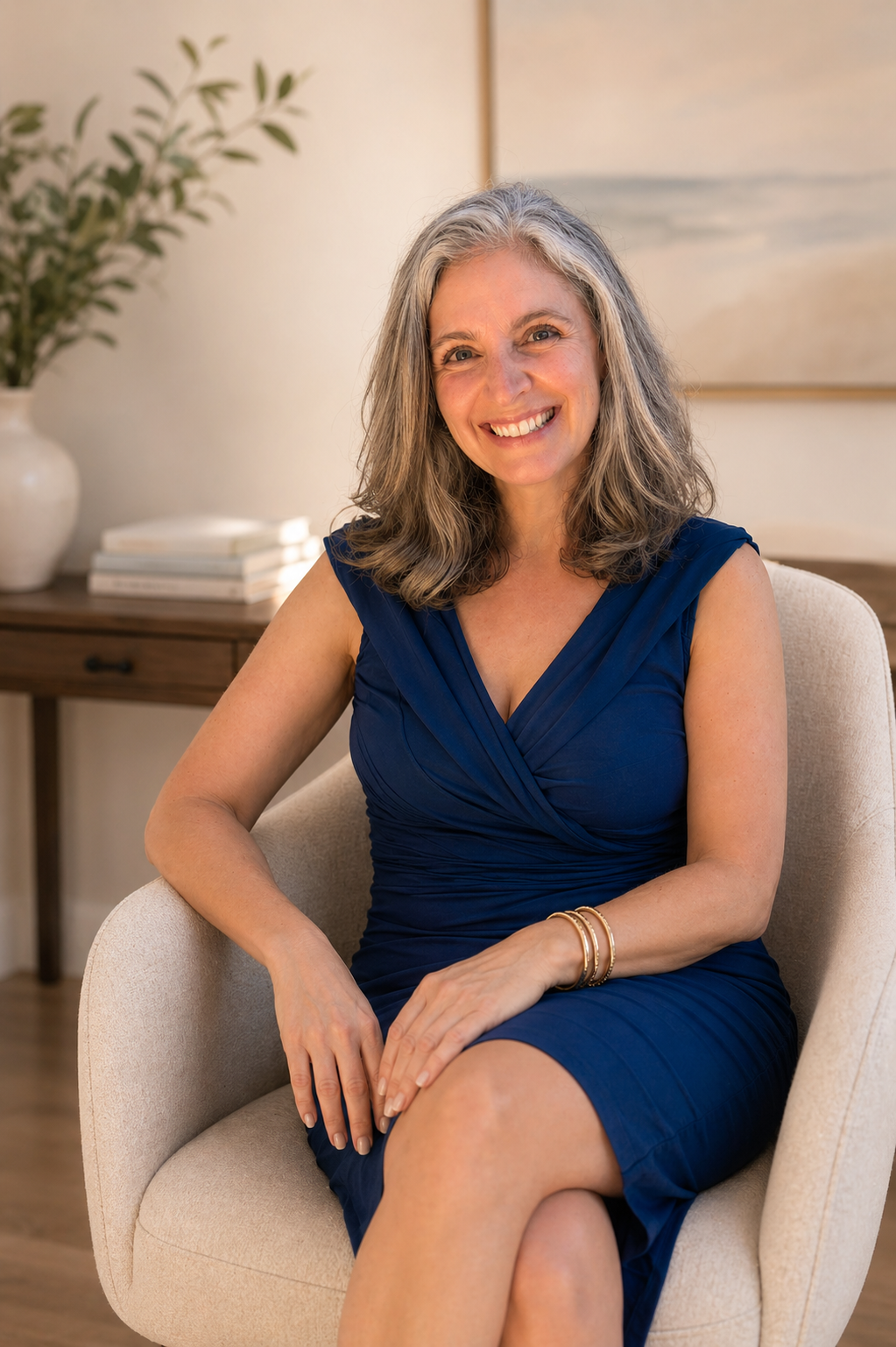 A middle-aged woman with gray hair, wearing a sleeveless navy blue dress, seated in a beige armchair, smiling at the camera in a modern interior setting.