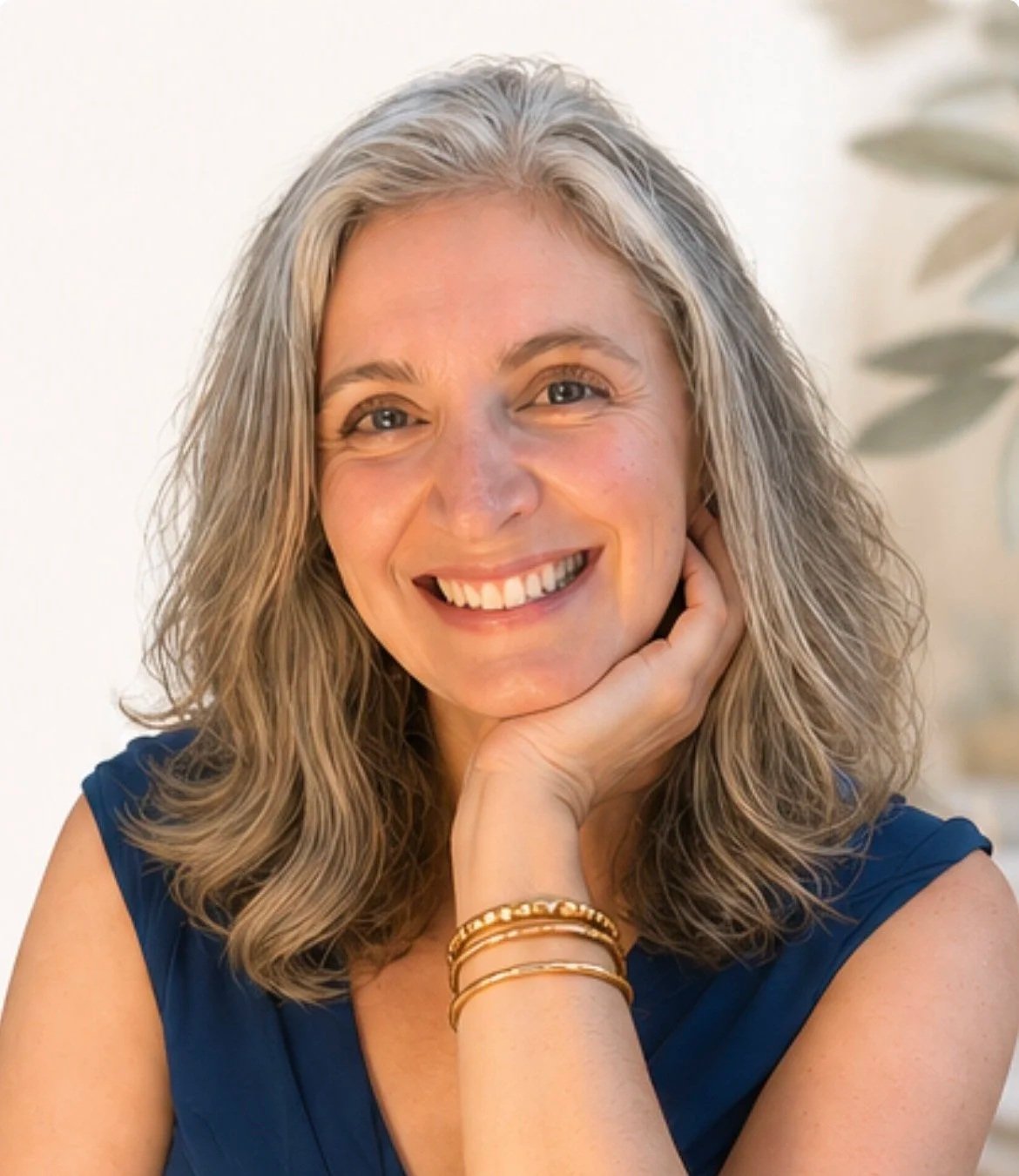 A woman, Dr. Bridget Colacchio, with wavy grey hair smiling and resting her chin on her hand, wearing a blue sleeveless top and gold bracelets, in a bright room with a plant in the background.
