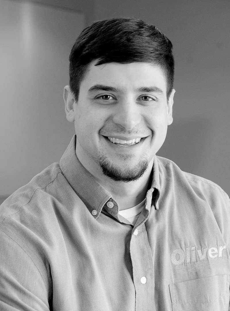 Young man with short dark hair and a beard, smiling, wearing a collared shirt with a logo, in a black and white portrait.
