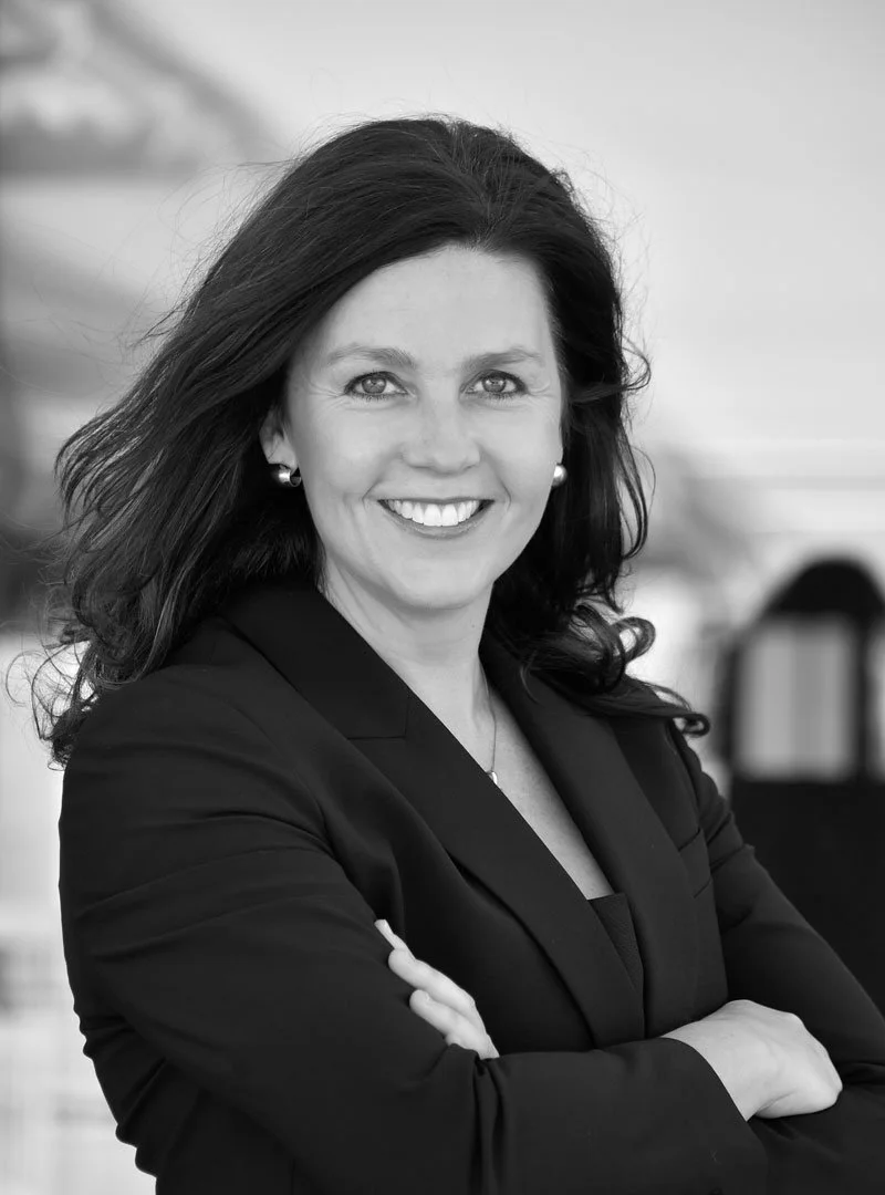 Black and white portrait of a smiling woman with dark, shoulder-length hair, wearing a black blazer and pearl earrings, standing with arms crossed in a professional setting.