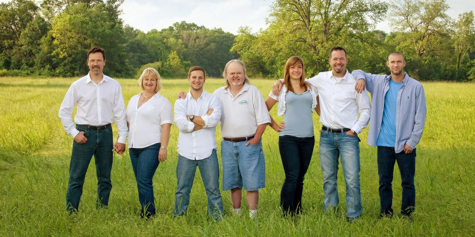Group of eight people standing arm-in-arm in a grassy field with trees in the background, smiling