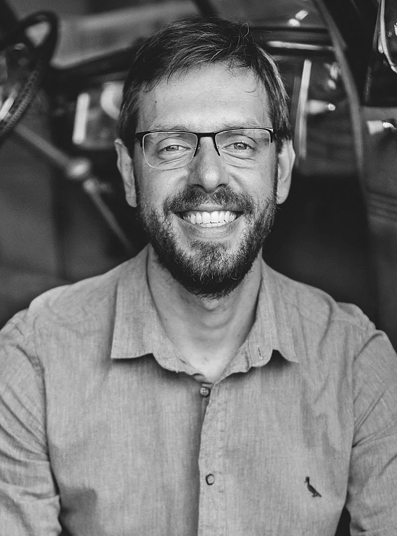 A smiling man with glasses, a beard, and a button-up shirt, sitting in an indoor setting with shelves and various objects in the background.