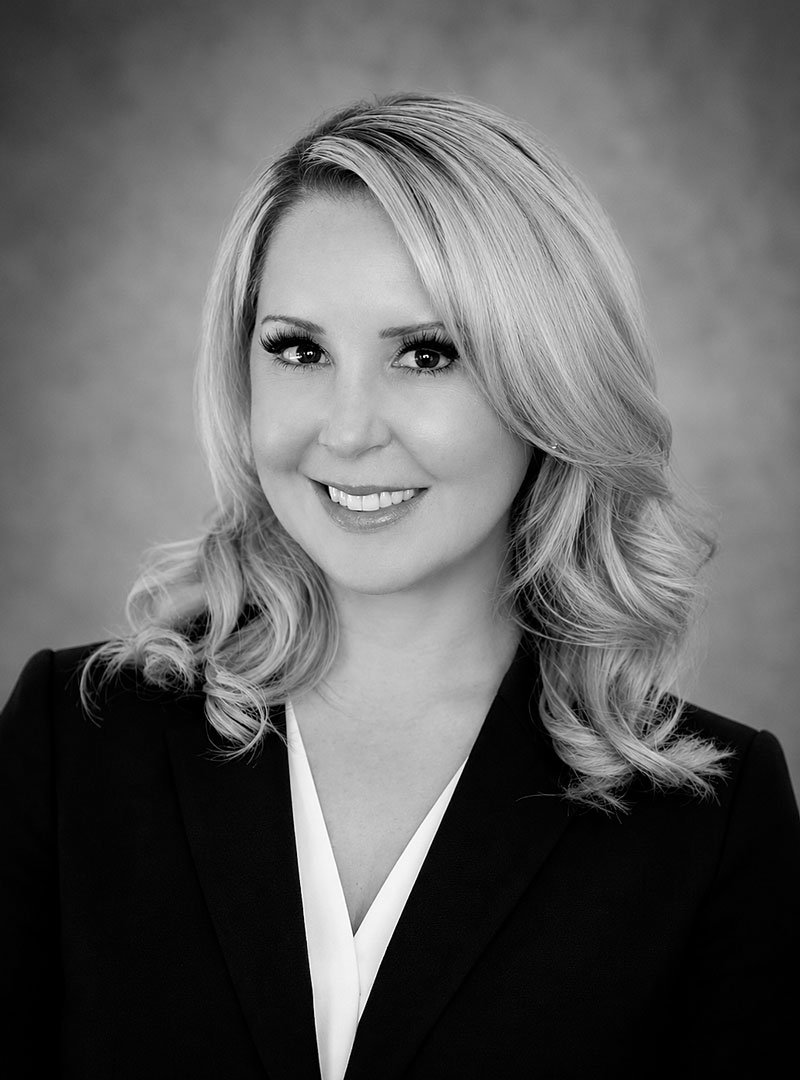 Black and white professional headshot of a woman with shoulder-length wavy blonde hair, smiling, wearing a dark blazer and a light blouse.