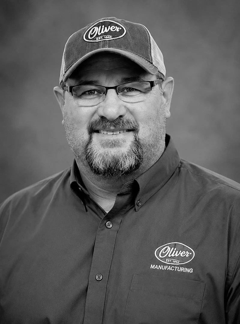 A smiling man with glasses, a beard, and a cap that says 'Oliver' wearing a collared shirt with an 'Oliver Manufacturing' logo.