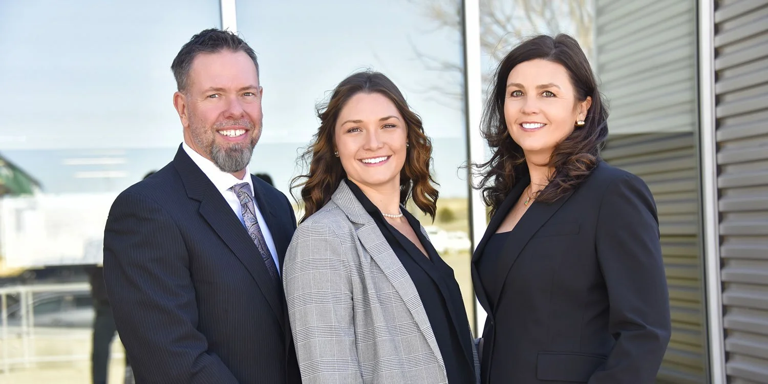Three professionally dressed people smiling outdoors in front of a modern building with glass and metal siding.