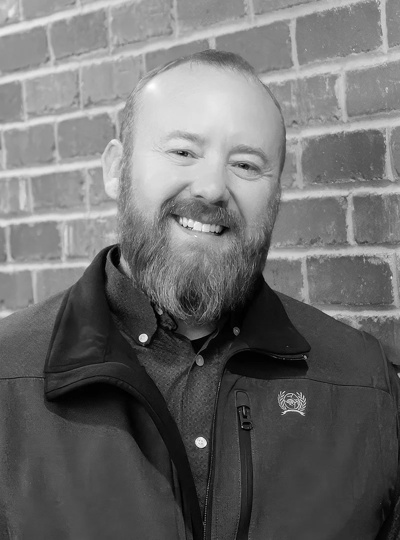 A smiling man with a beard and short hair, wearing a dark jacket and button-up shirt, standing in front of a brick wall.