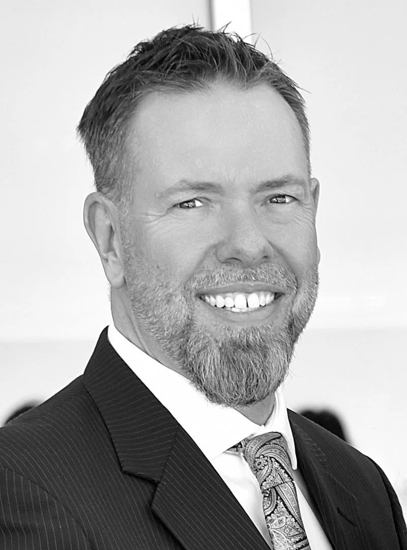 Black and white portrait of a smiling man with short hair, a beard, wearing a suit and tie.
